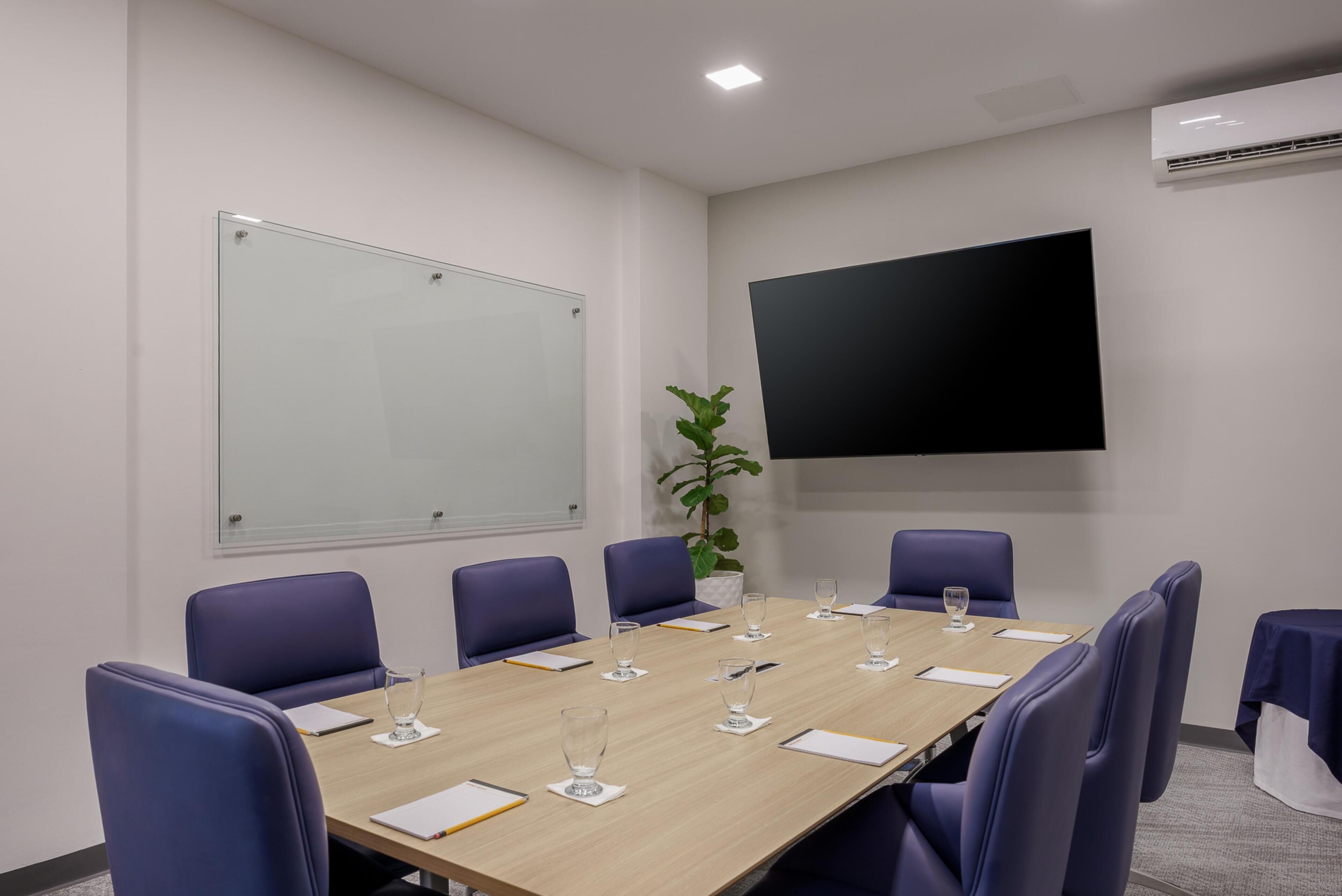 Una sala de conferencias con una larga mesa, sillas azules, una pizarra y un televisor de pantalla plana.