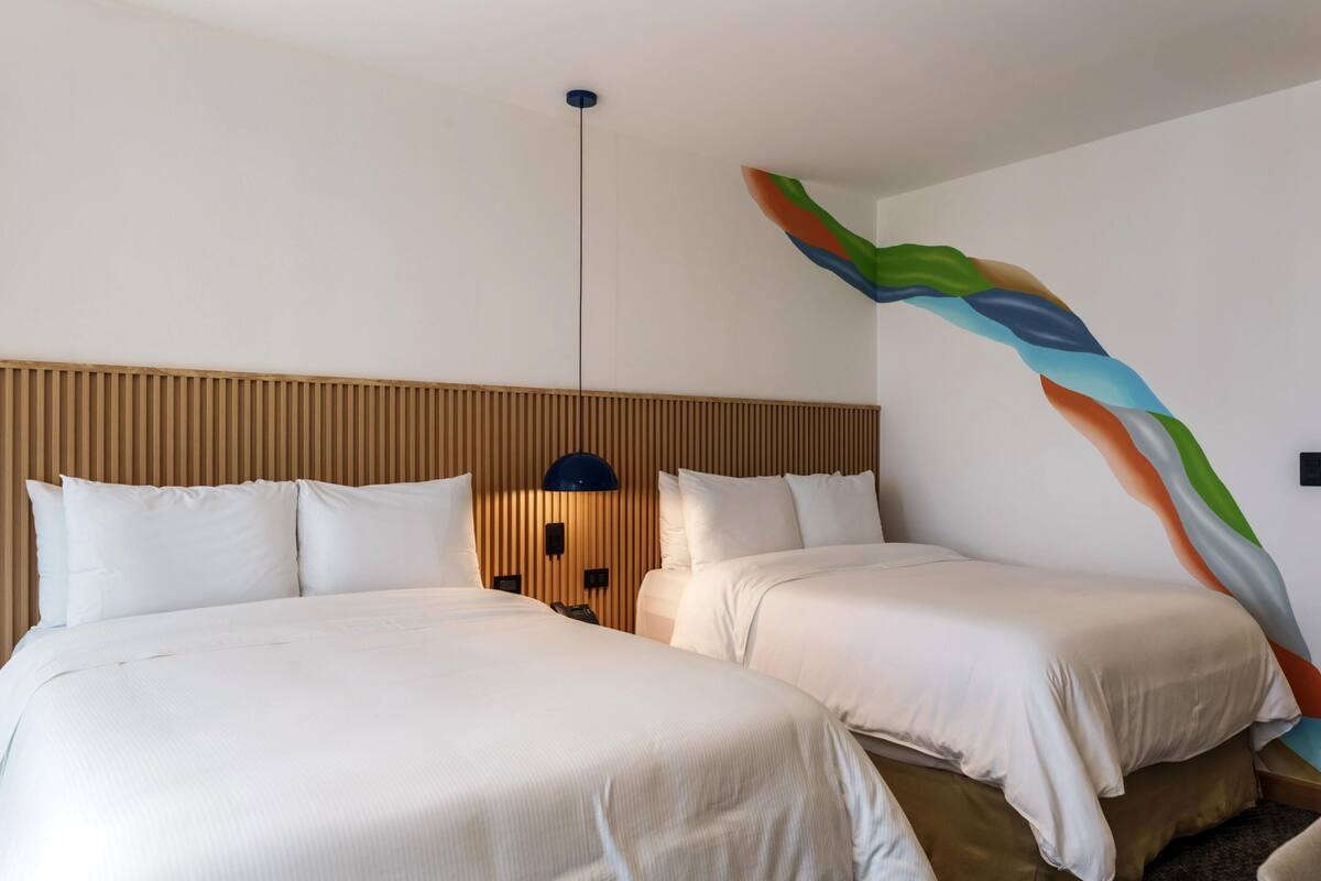 Quarto de hotel com duas camas, uma cabeceira de madeira, uma luminária fixada na parede e uma pintura abstrata colorida.