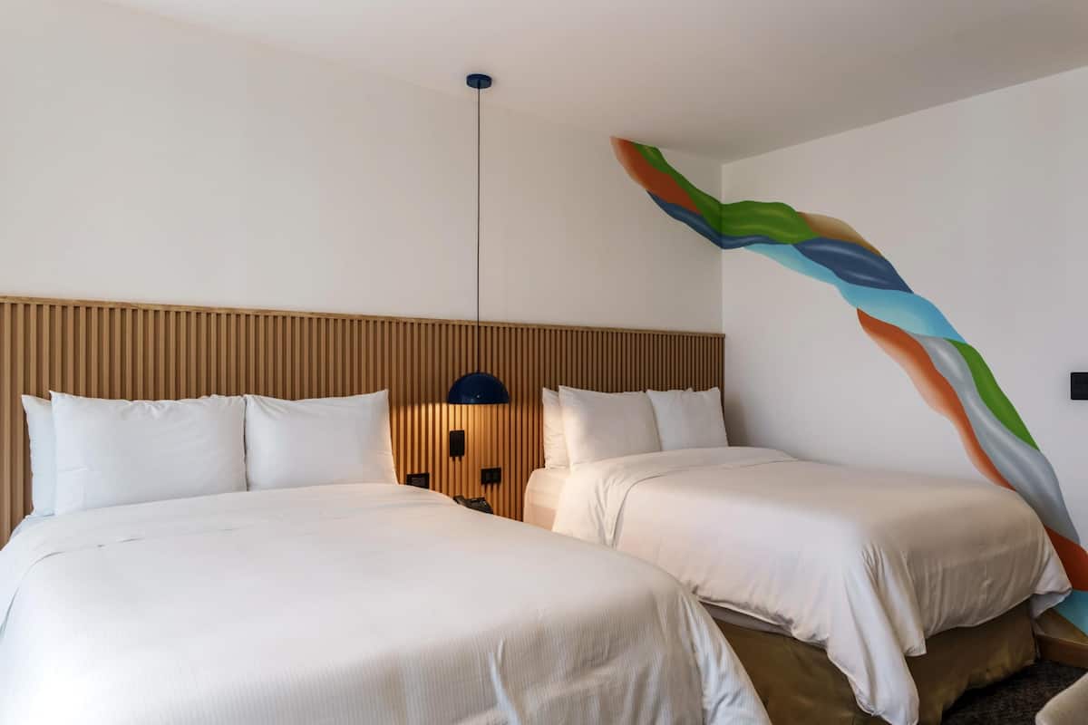 Une chambre d’hôtel avec deux lits, une tête de lit en bois, une lampe fixée au mur et un tableau abstrait aux couleurs vives.