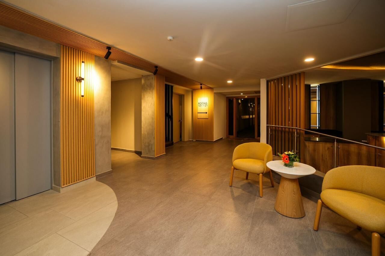 Un hall d’hôtel comprenant un comptoir d’accueil, un espace salon avec deux fauteuils et un panneau indiquant les numéros de chambre 101 à 121.