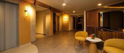 Un hall d’hôtel comprenant un comptoir d’accueil, un espace salon avec deux fauteuils et un panneau indiquant les numéros de chambre 101 à 121.