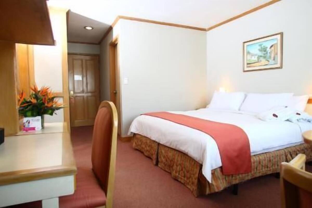 Quarto de hotel com cama, escrivaninha, cadeira, porta e um quadro na parede.