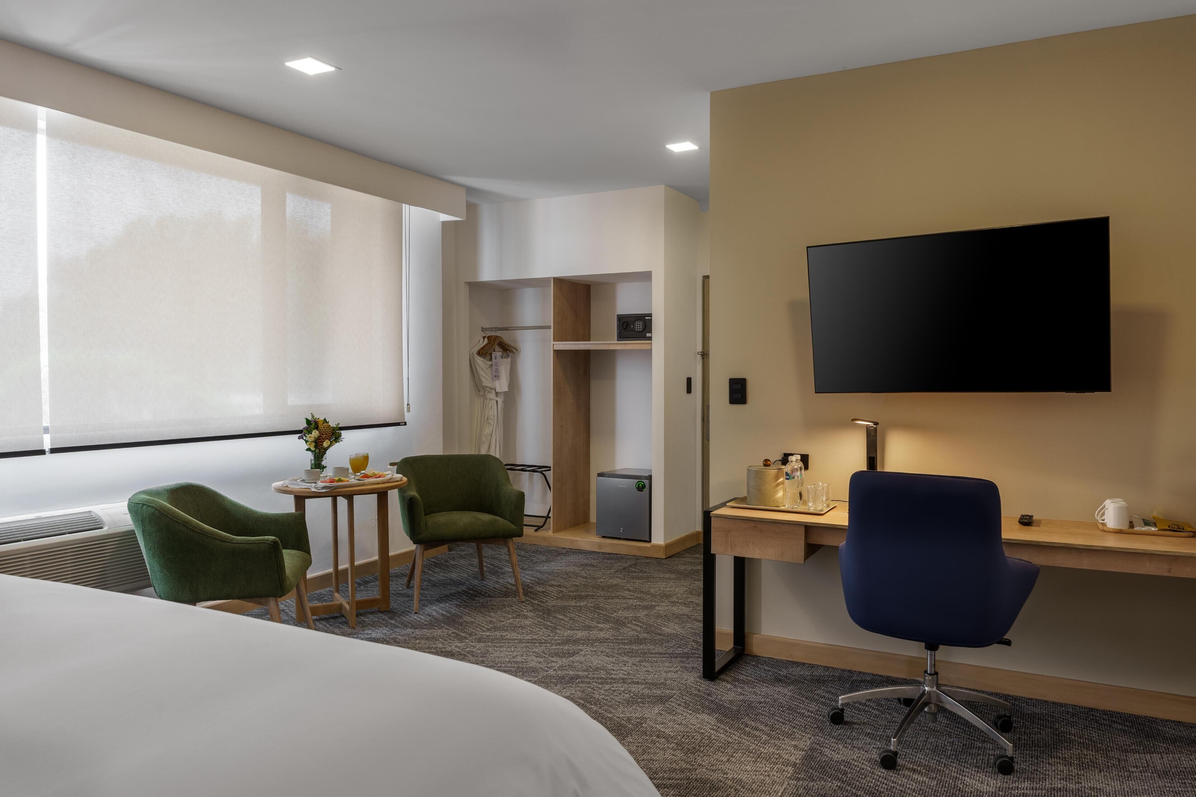 A hotel room with a bed, a desk with a chair, a TV, a small table with a chair, a closet, and a window.