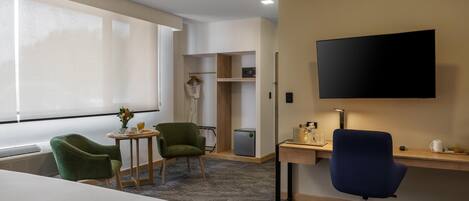 A hotel room with a bed, a desk with a chair, a TV, a small table with a chair, a closet, and a window.