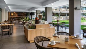 A modern restaurant with wooden tables and chairs, a central bar area with a fruit display, and large windows offering a view of the outdoor garden. - Best Western Plus Hotel Terraza (San Salvador)