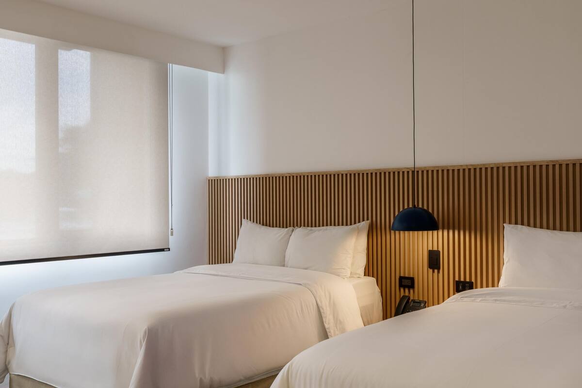 Quarto de hotel com duas camas, uma cabeceira de madeira, mesa de cabeceira com abajur e uma janela grande.