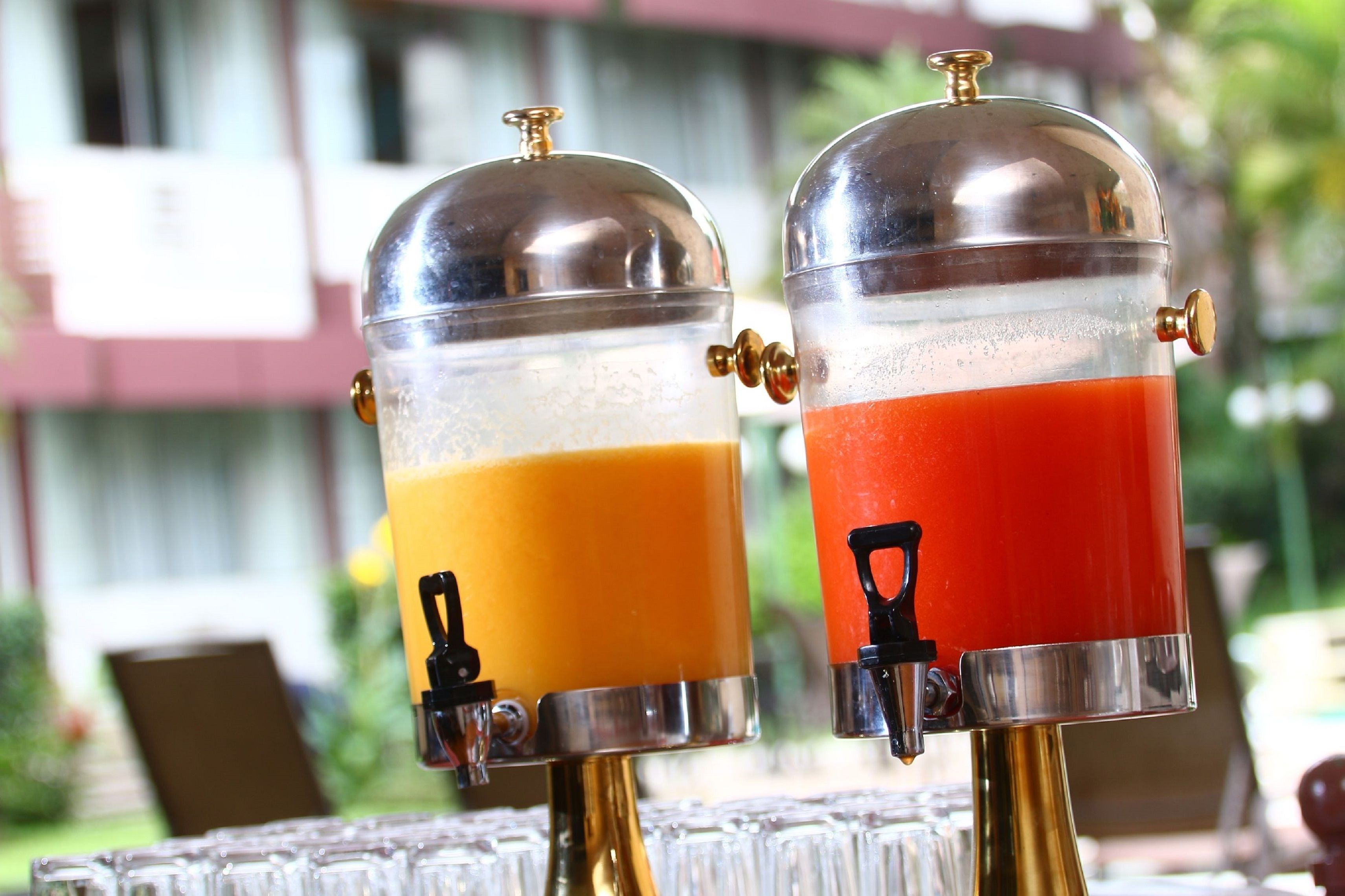 Two beverage dispensers with transparent sections, one containing orange juice and the other red juice, placed outdoors with a building in the background.