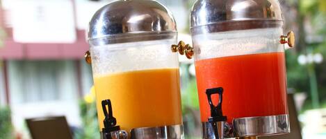 Two beverage dispensers with transparent sections, one containing orange juice and the other red juice, placed outdoors with a building in the background.