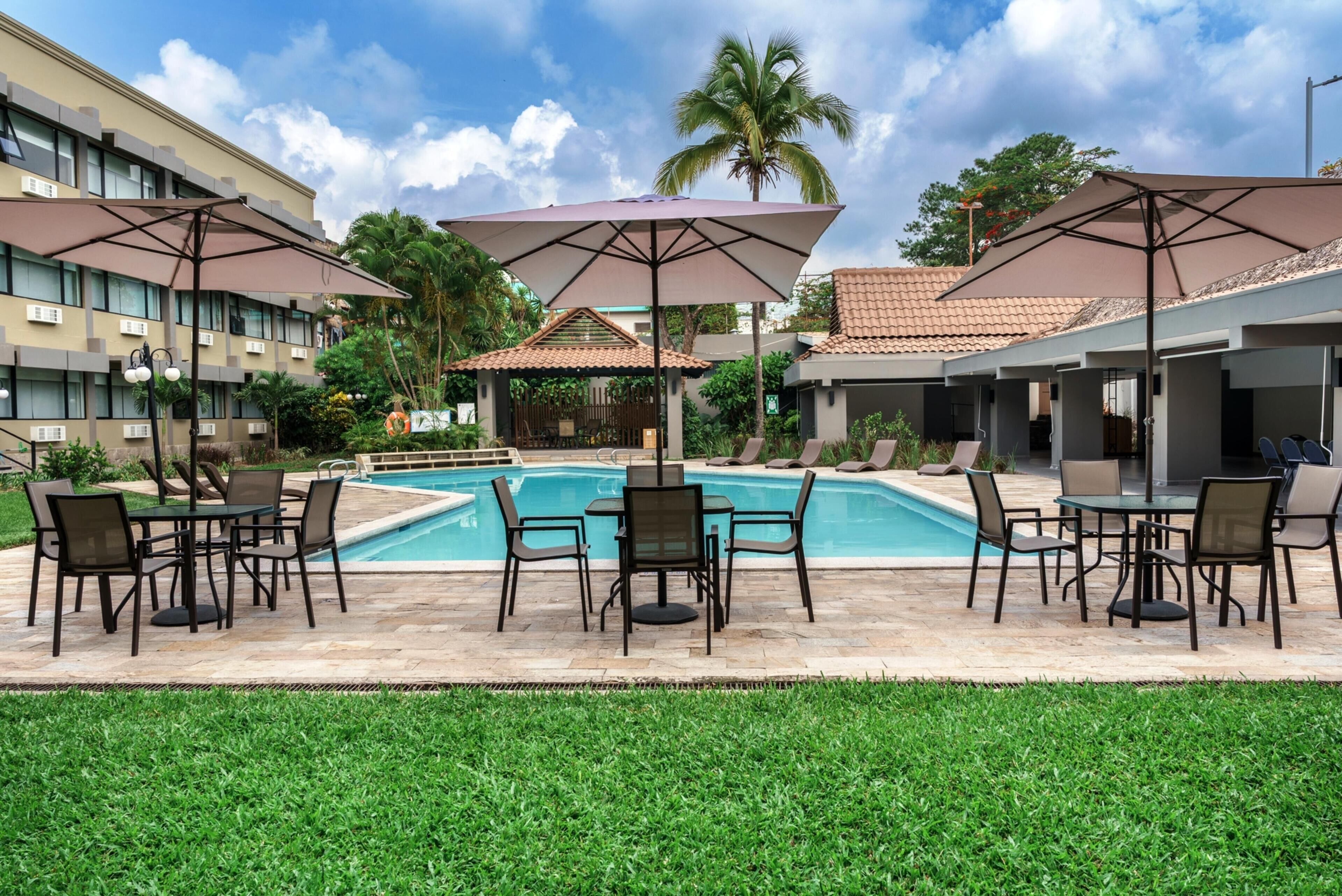 A poolside area with outdoor seating and umbrellas, surrounded by buildings and palm trees.