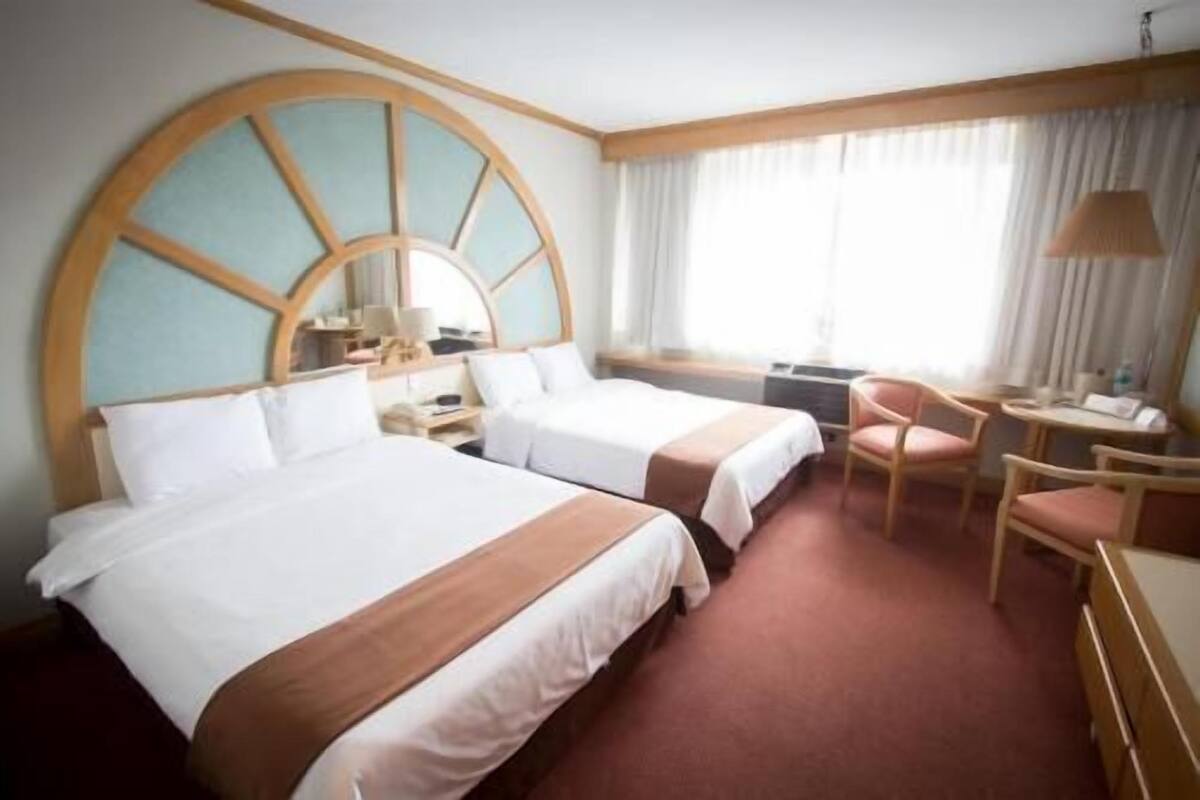 Quarto de hotel com duas camas, uma escrivaninha, uma cadeira e uma janela grande com cortinas.