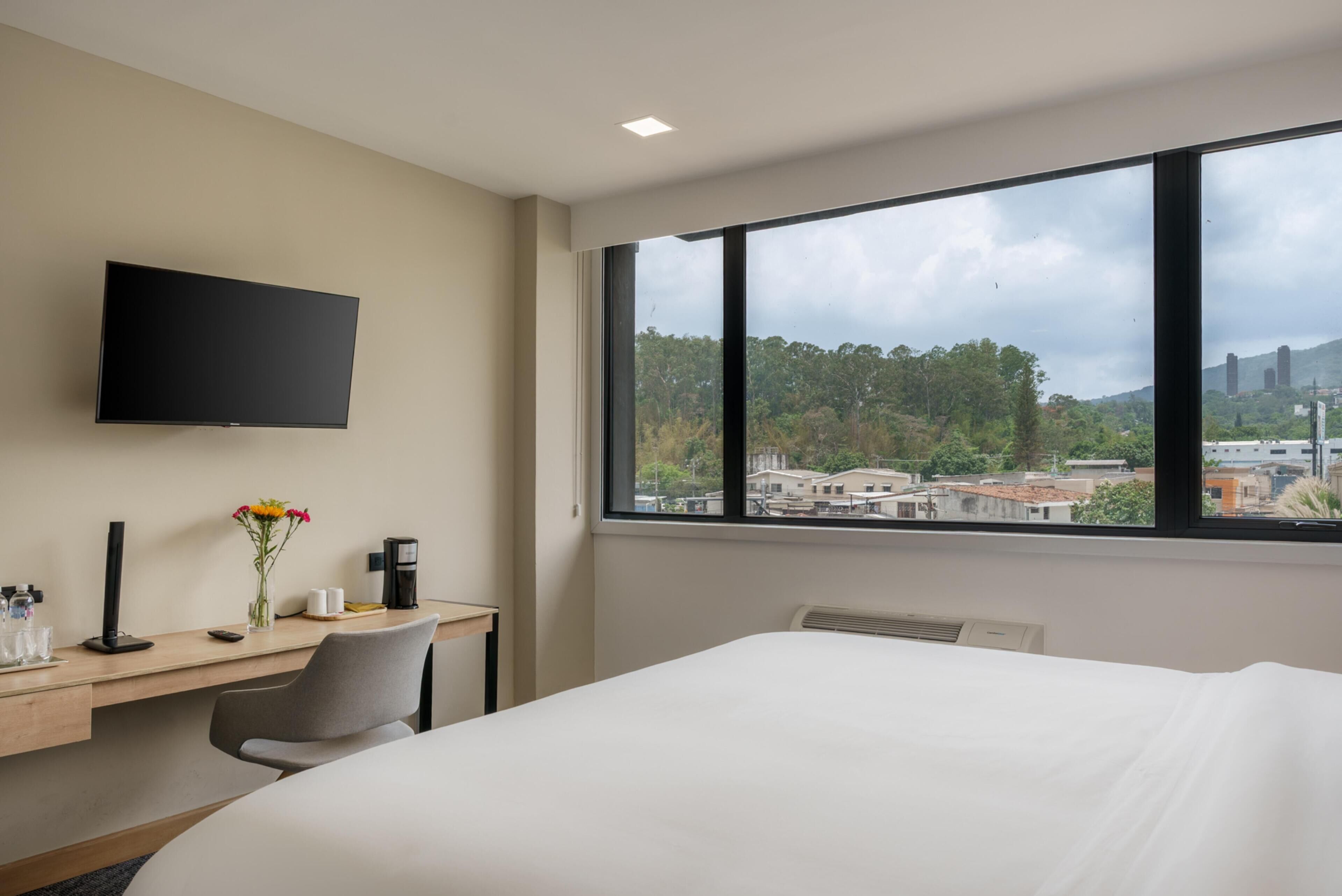 A hotel room with a bed, a desk with a chair, a flat-screen TV, and a large window offering a city view.