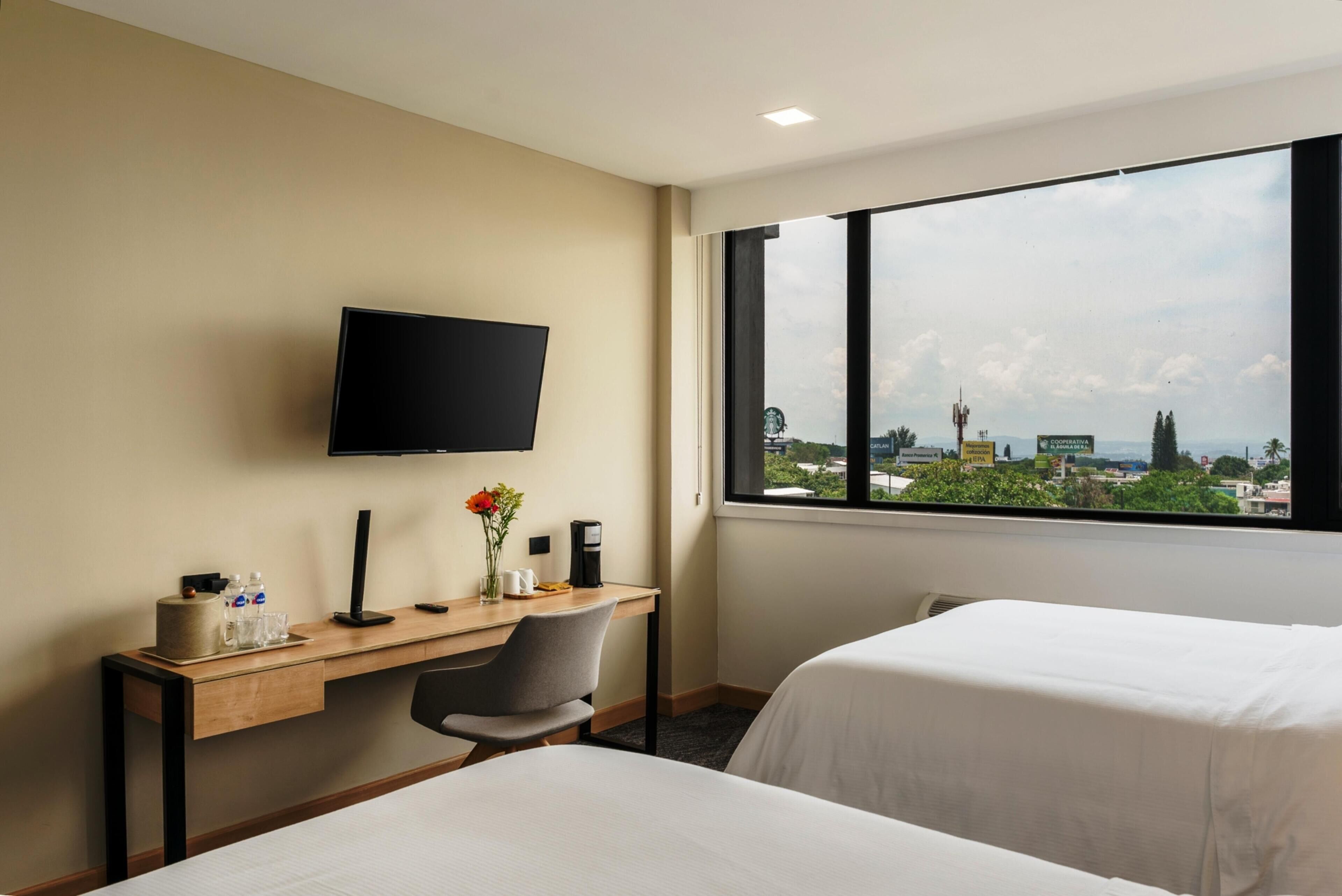 A hotel room with a desk, a TV mounted on the wall, two beds, and a large window offering a city view.