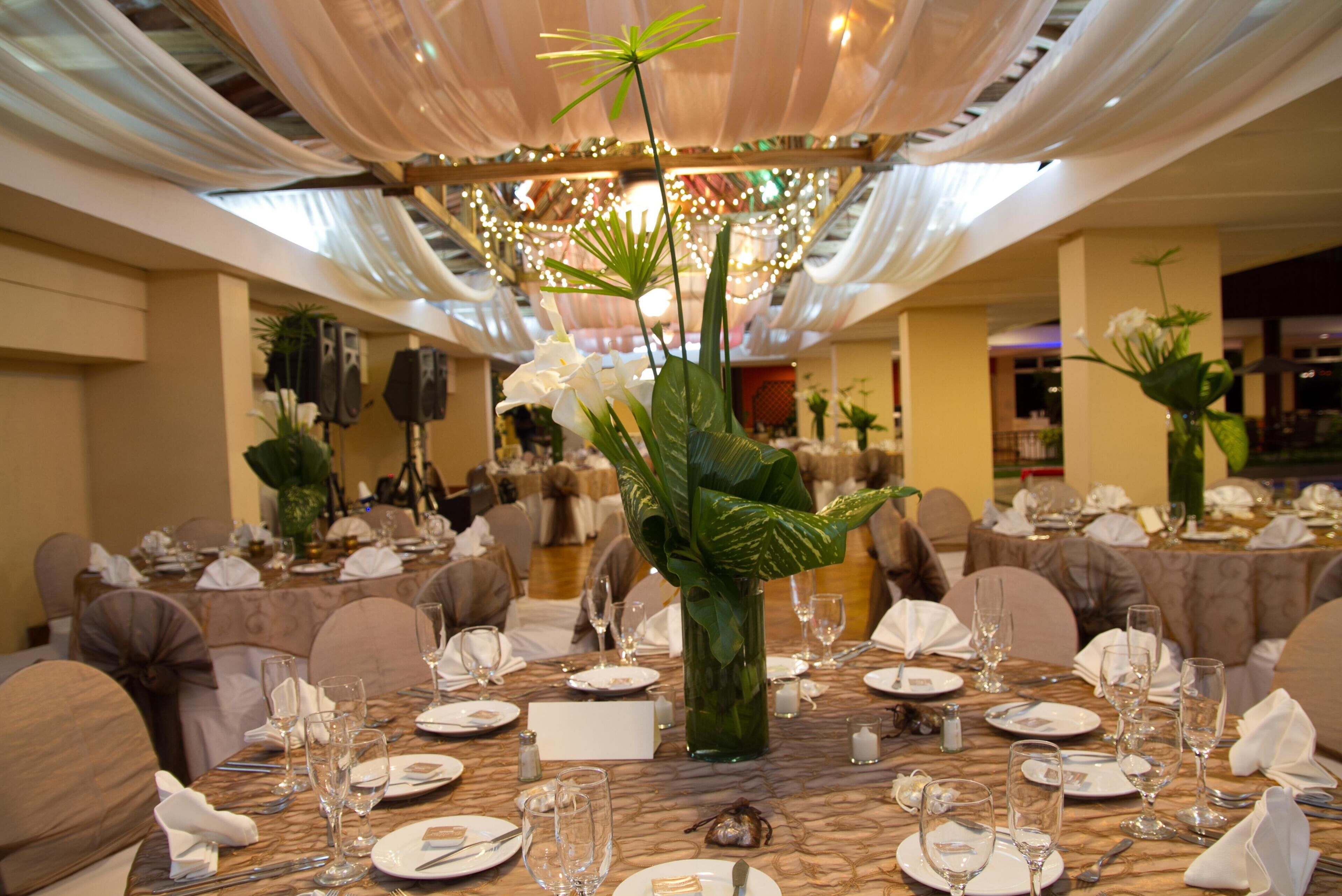 Un salón de banquetes con mesas redondas dispuestas para cenar, una gran lámpara de araña y plantas decorativas.