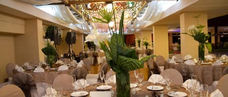 Un salón de banquetes con mesas redondas dispuestas para cenar, una gran lámpara de araña y plantas decorativas.