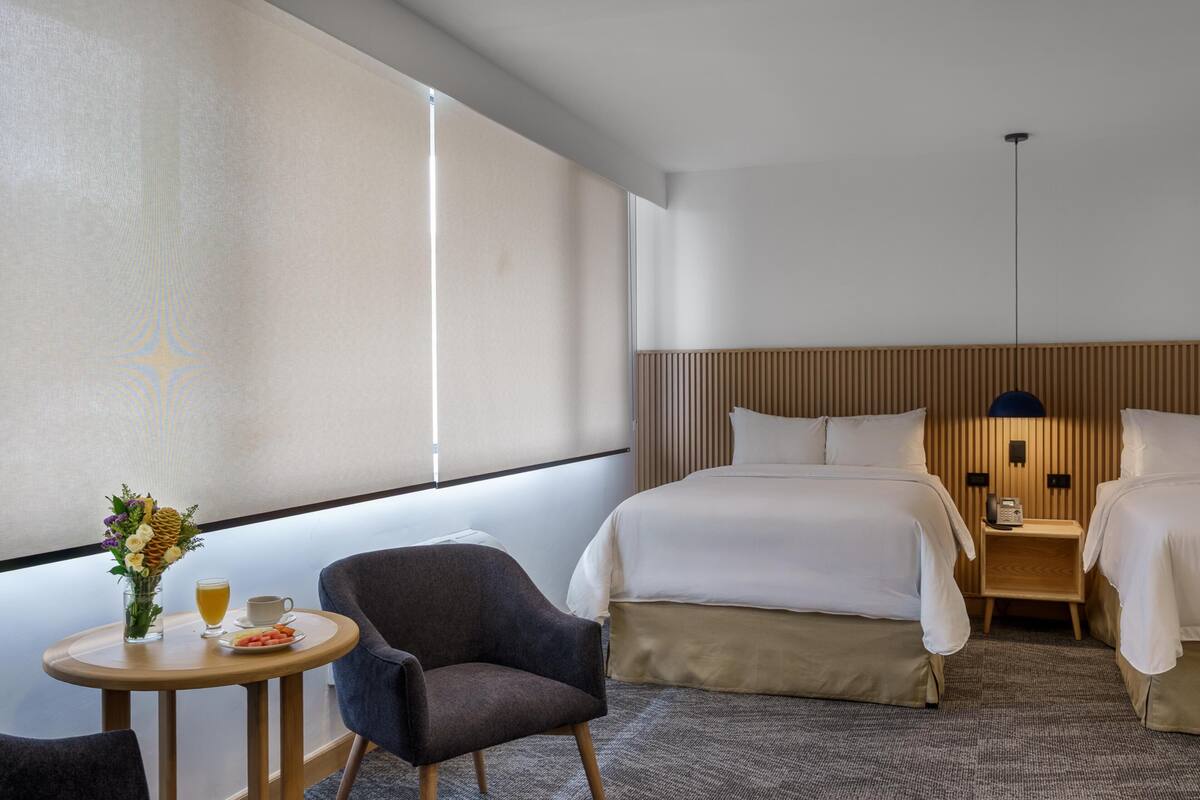 Quarto de hotel com uma cama, mesas de cabeceira, uma cadeira, uma mesinha com bebida e fruta, e uma janela com persianas.