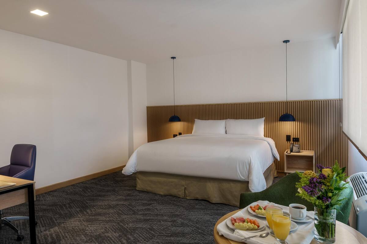Quarto de hotel com cama, mesas de cabeceira, escrivaninha, cadeira e bandeja de café da manhã com alimentos e bebidas.