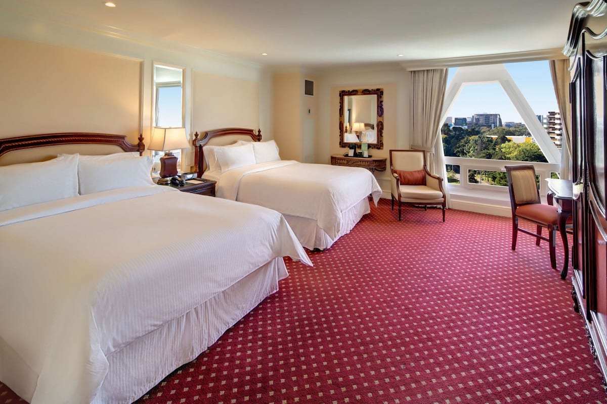 A hotel room with two beds, a sitting area with a chair, a small table, and a view of the city.