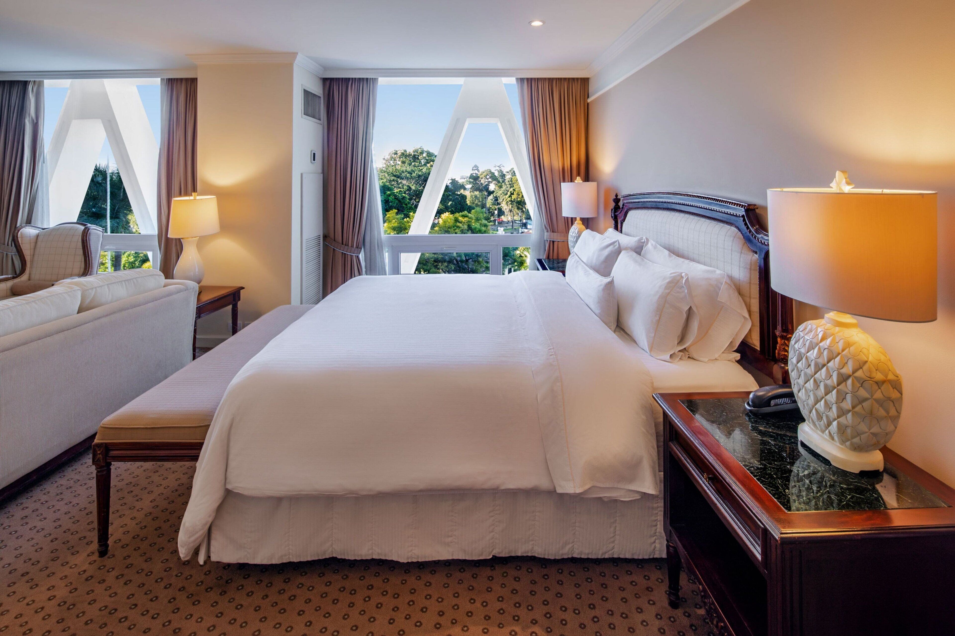 Una habitación de hotel con una cama grande, dos mesitas de noche, una silla y vistas a zonas verdes.