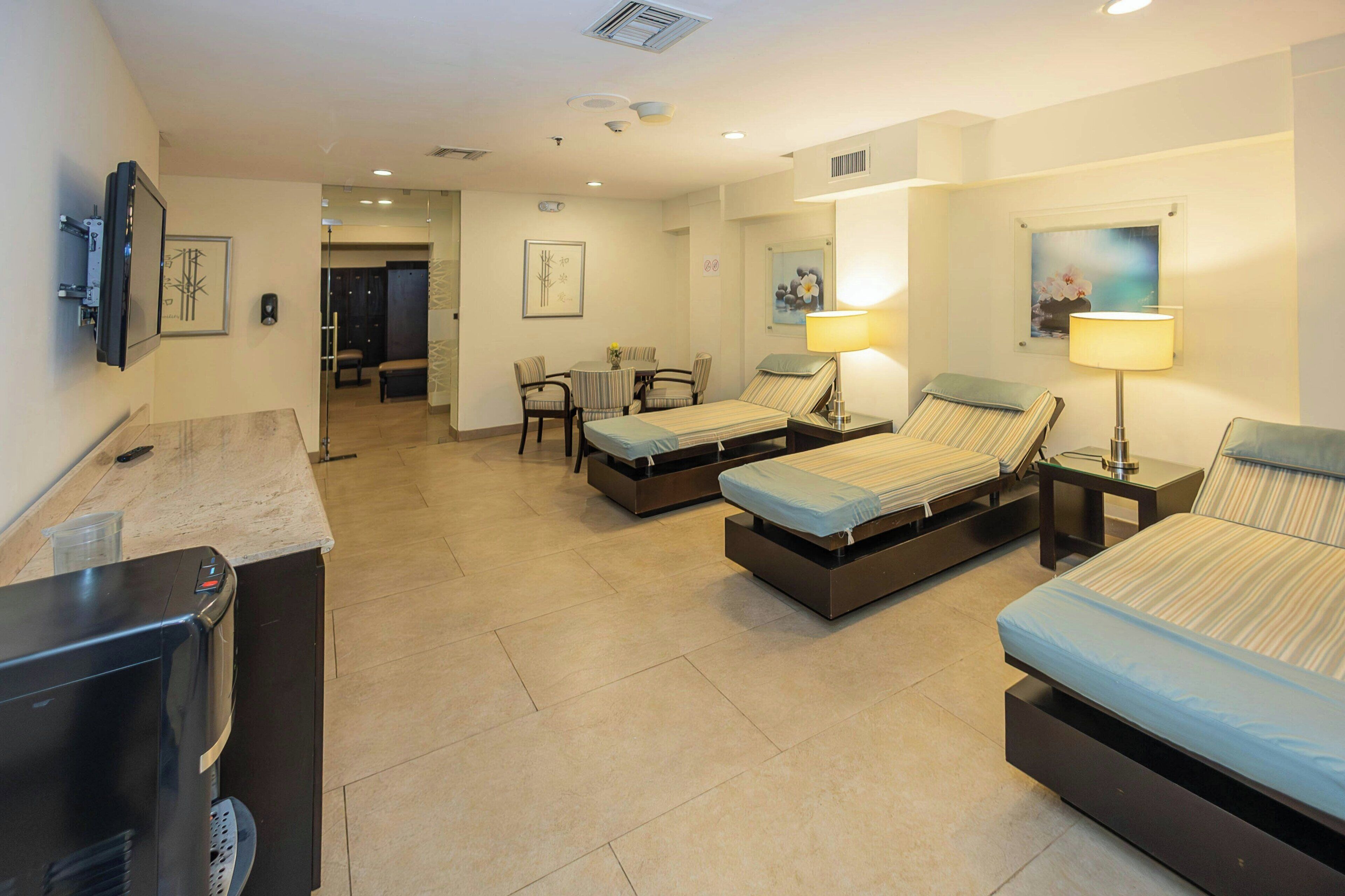 A hotel room with two single beds, a desk with a computer, a chair, a small table with a lamp, and a TV.