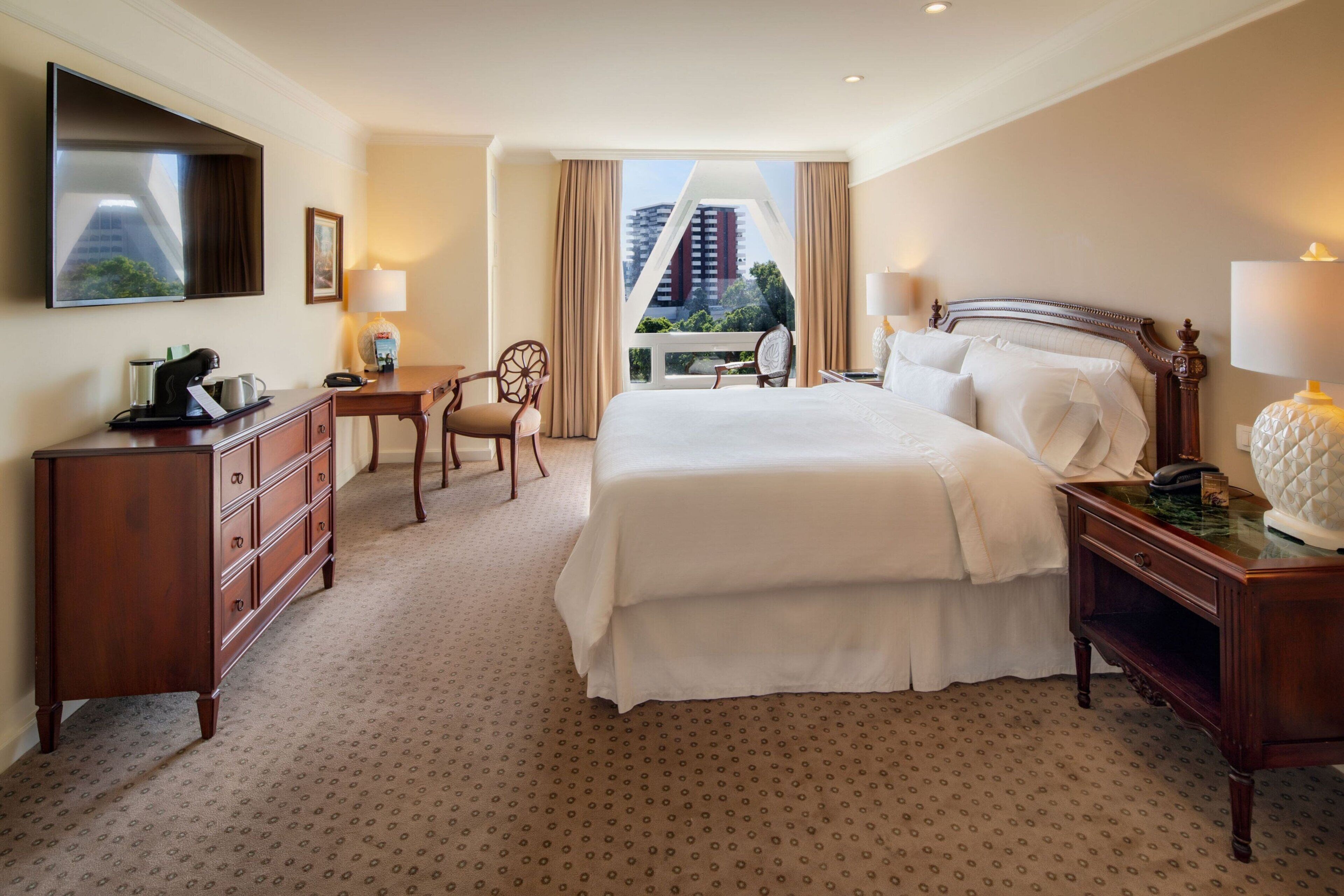 Une chambre d’hôtel avec un grand lit, un bureau avec une chaise, un téléviseur fixé au mur et une vue sur la ville par la fenêtre.