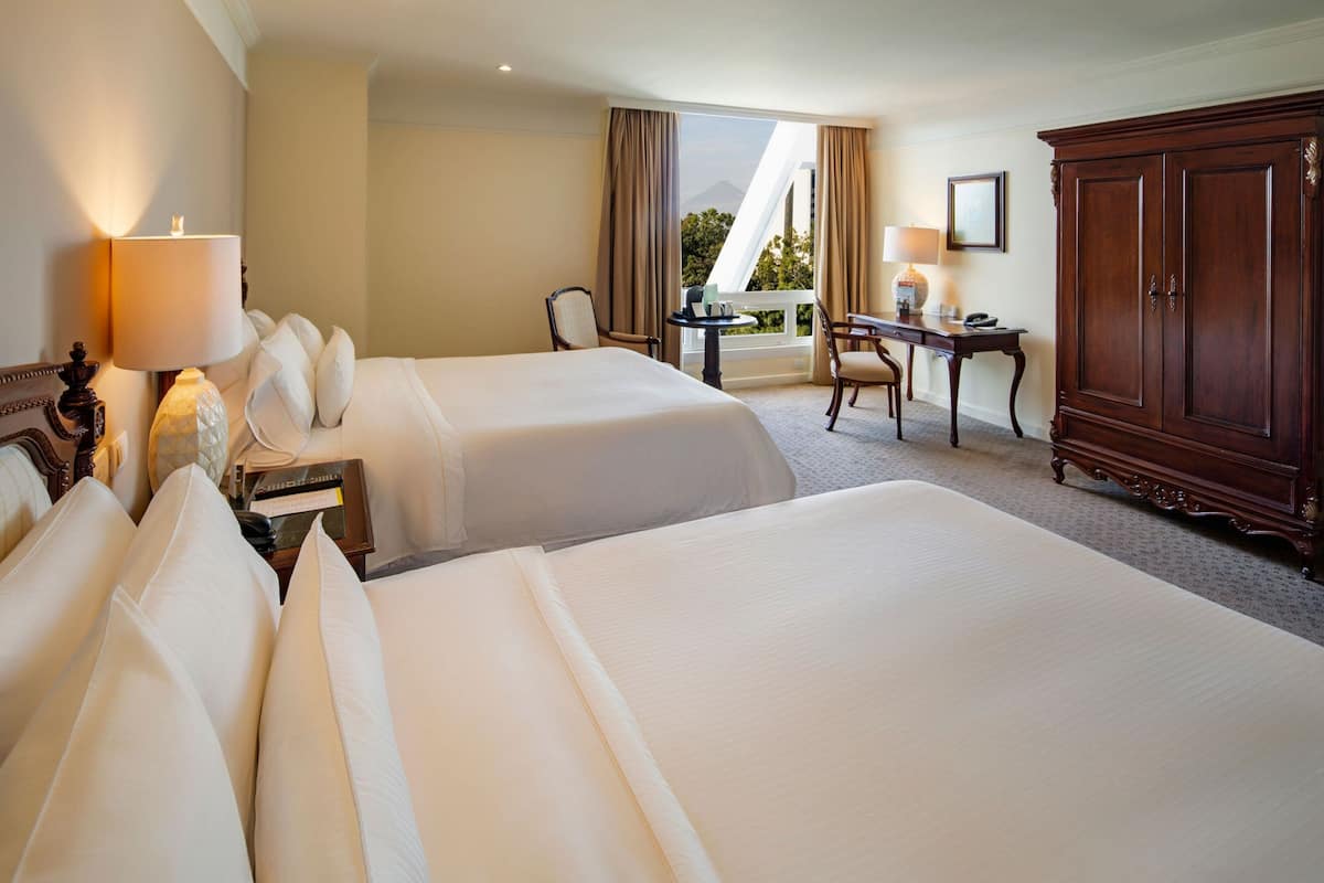 A hotel room with two beds, a desk, a chair, a lamp, and a large wooden cabinet.
