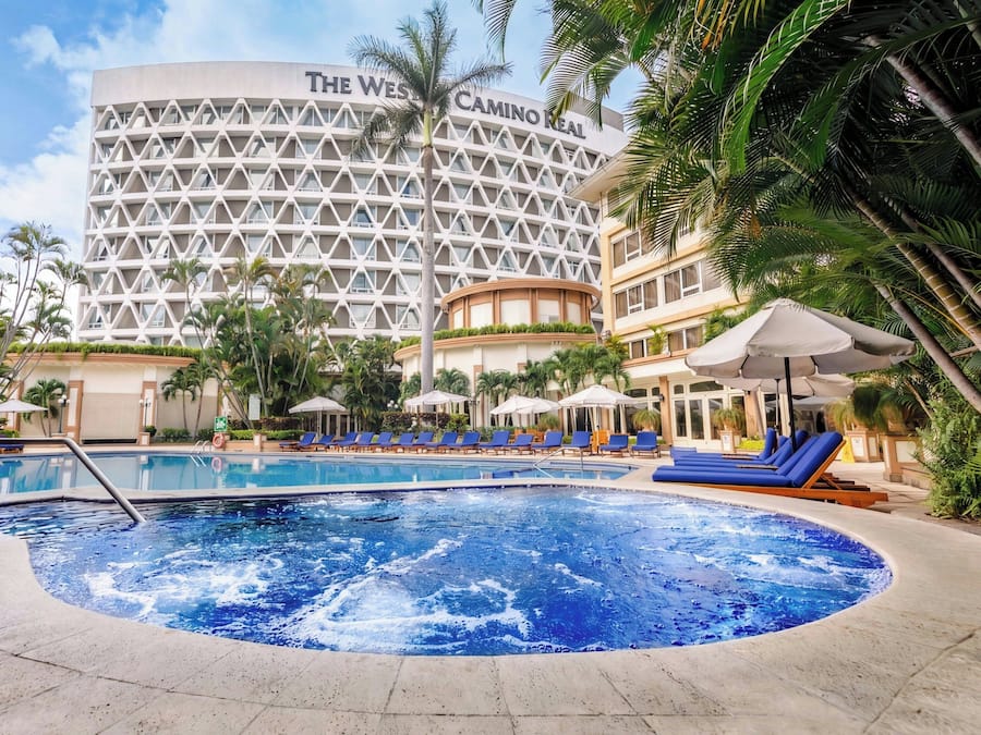 A hotel pool area with a large circular pool, lounge chairs, and palm trees.