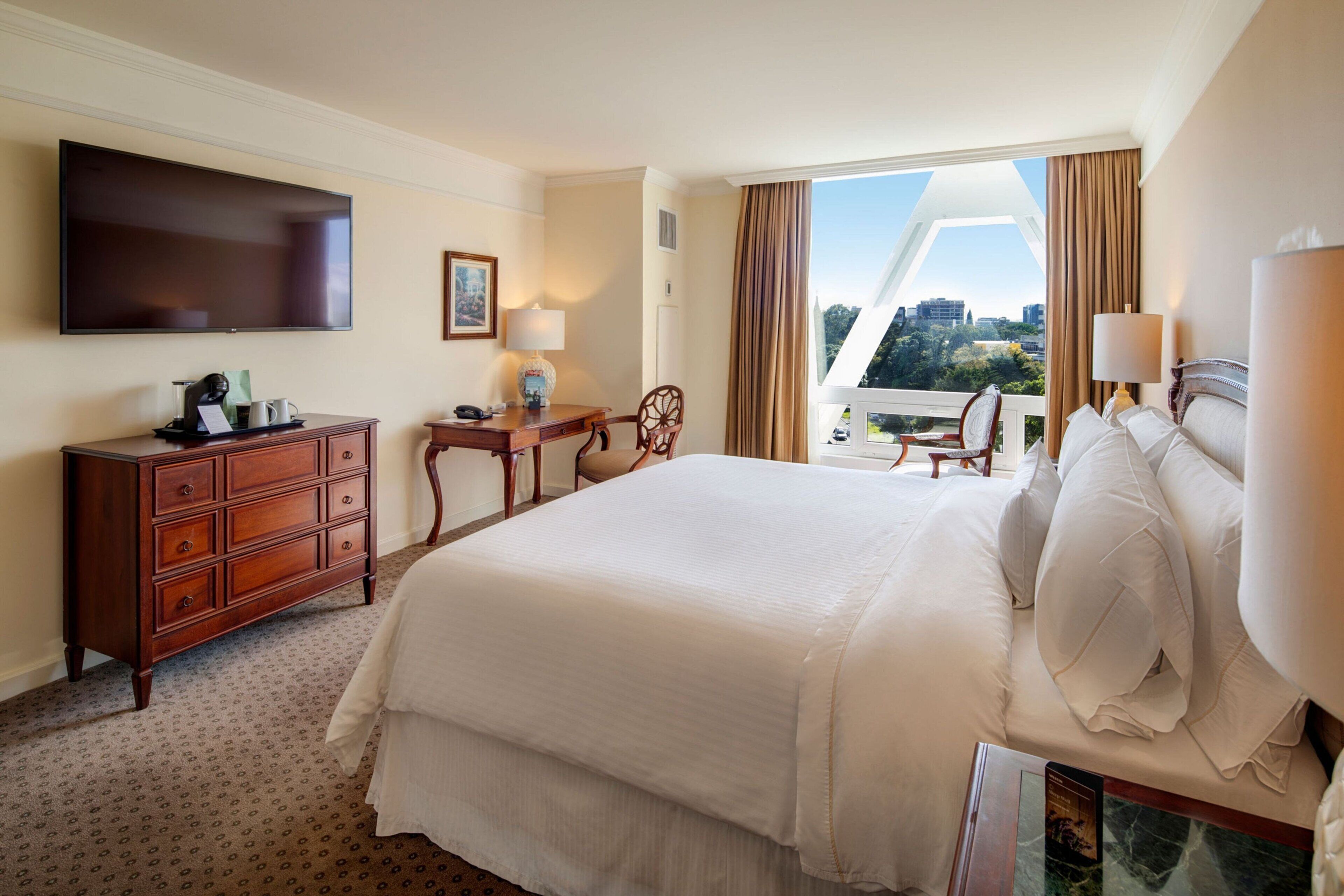 Een hotelkamer met een groot bed, een flatscreen televisie, een houten dressoir, een bureau met een lamp en uitzicht op de stad.