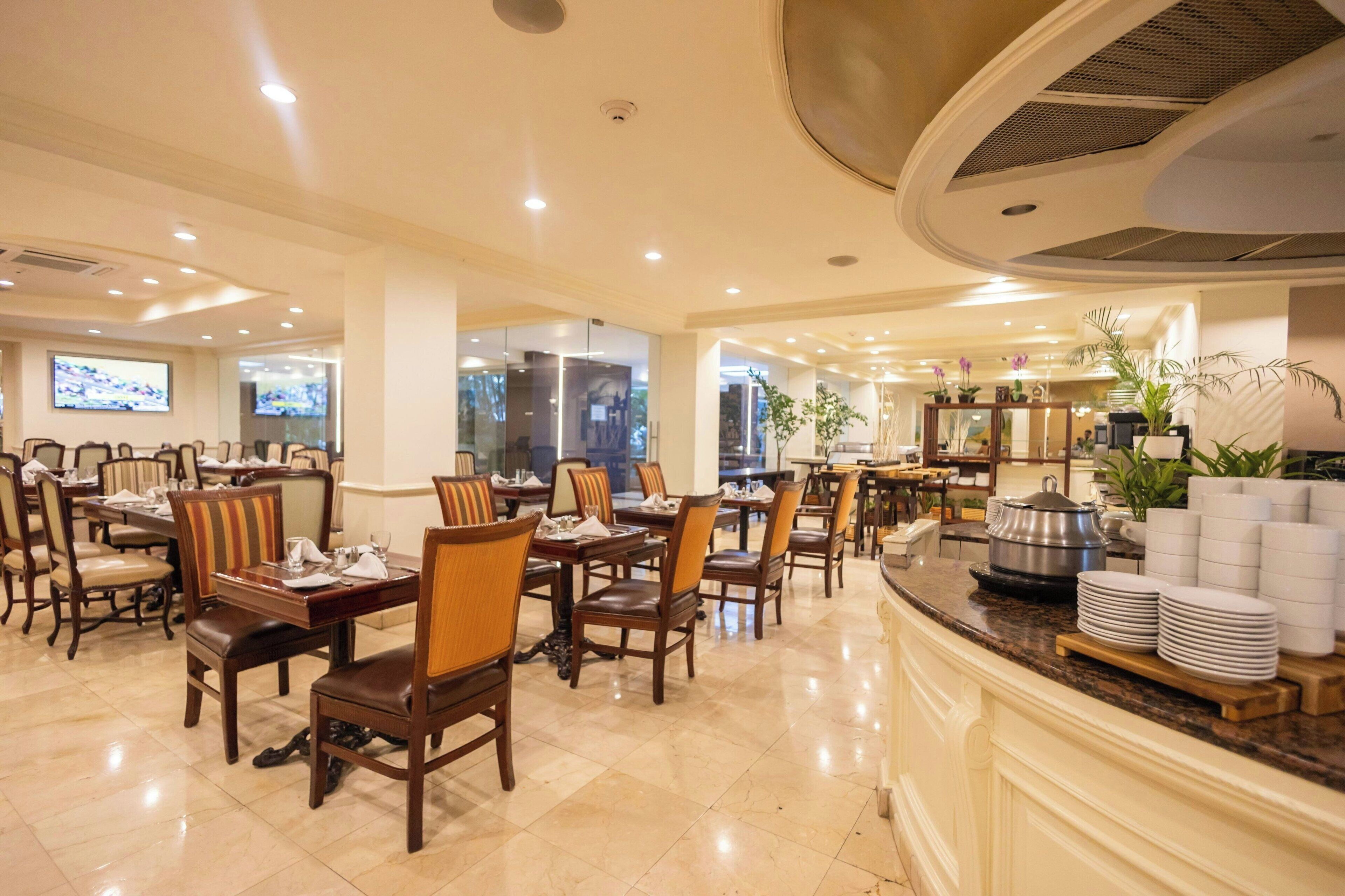 A hotel dining area with multiple tables and chairs, a buffet counter with plates and a pot, and a television screen in the background.