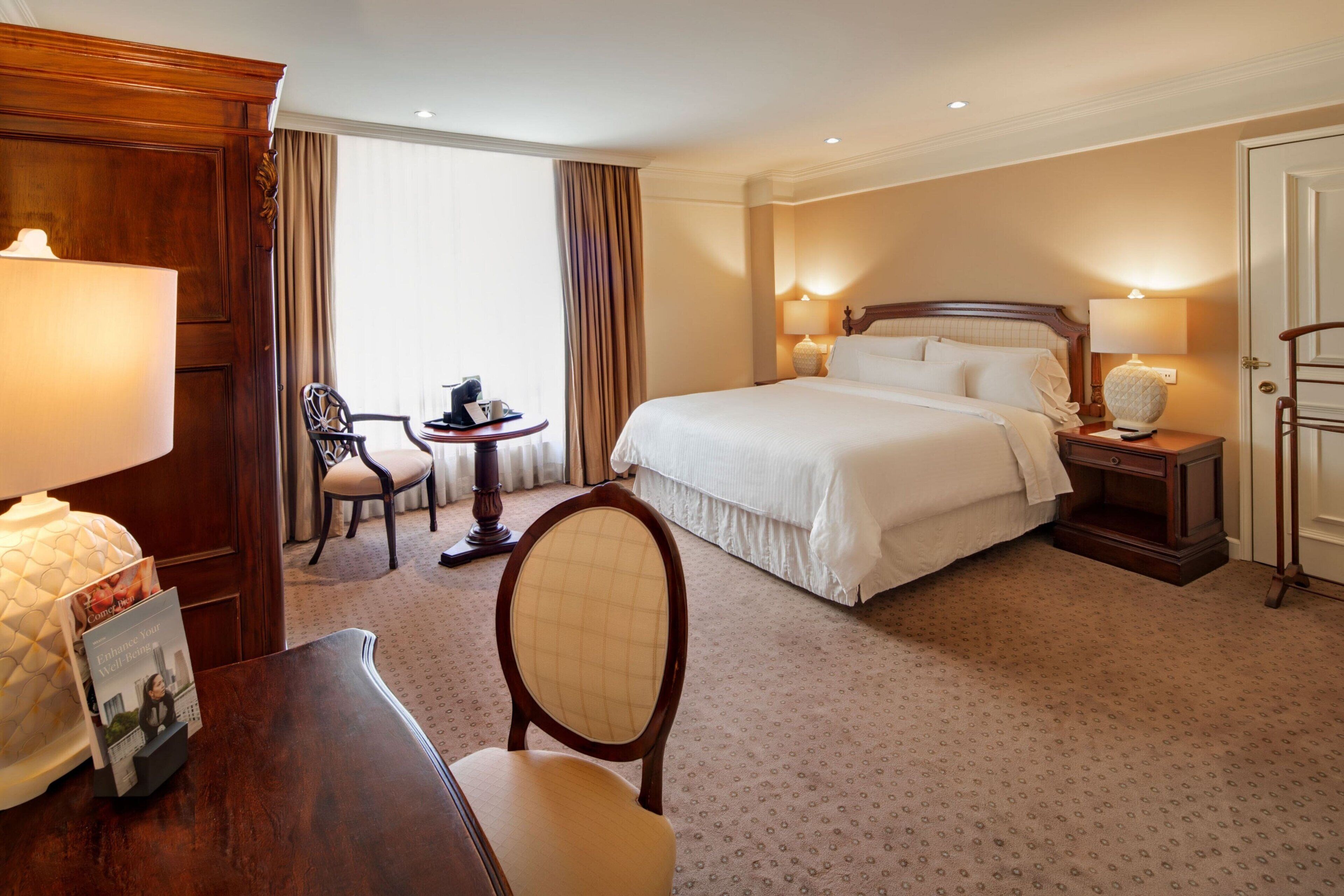 Quarto de hotel com uma cama grande, uma mesa com abajur, uma cadeira, uma mesinha com telefone e vista para o exterior através de uma janela.