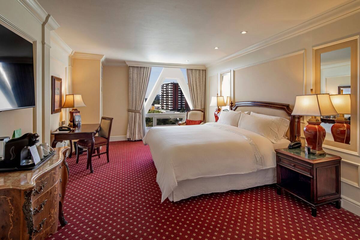 A hotel room with a large bed, two bedside tables with lamps, a desk with a chair, a dresser, and a large window with curtains.