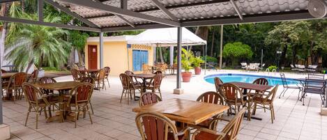 An outdoor patio area with wooden tables and wicker chairs, a pool, and a covered seating area with a white umbrella.