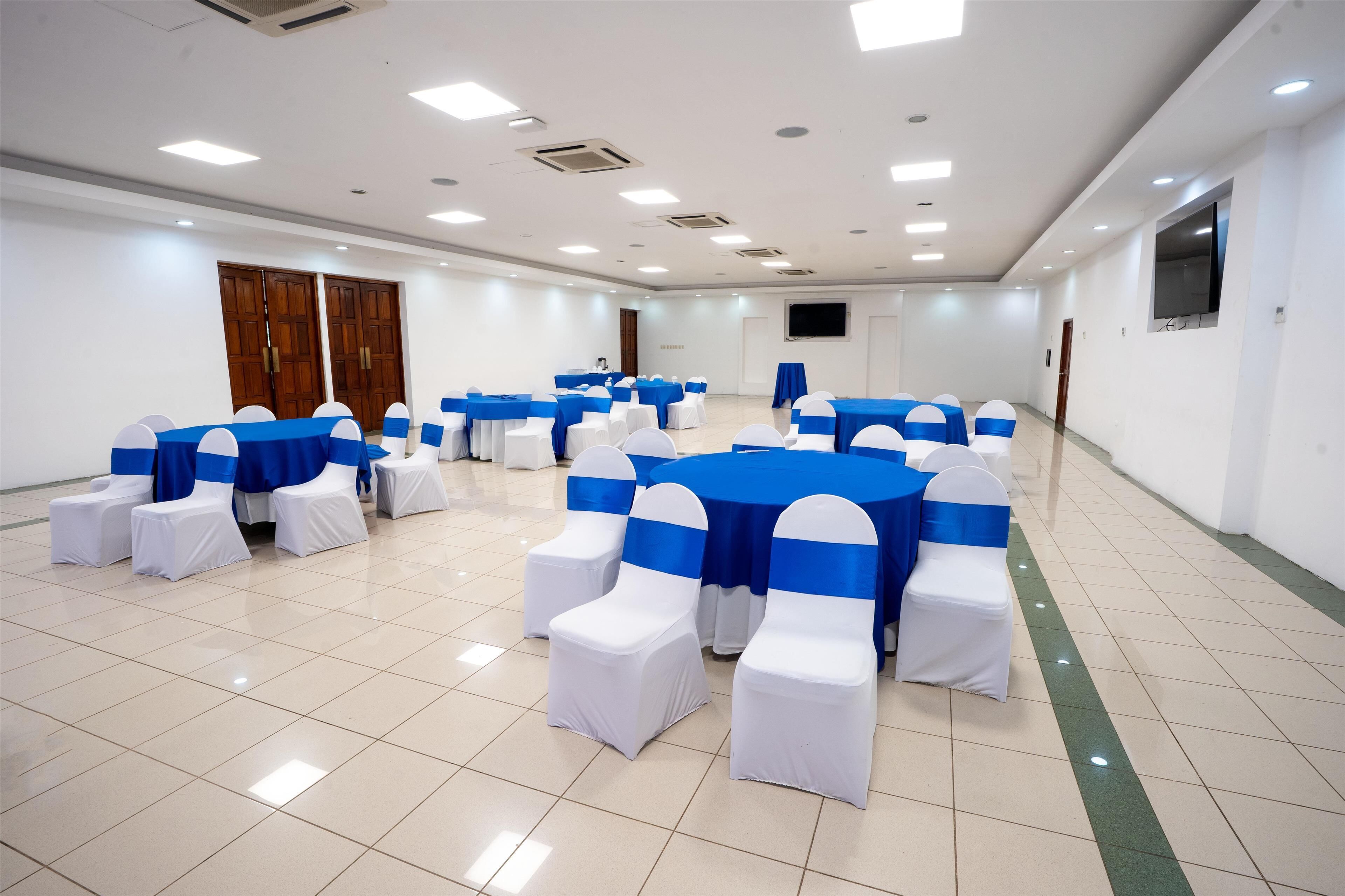 Un salón de conferencias con manteles blancos y azules, sillas blancas con cintas azules y una pantalla grande en la pared.