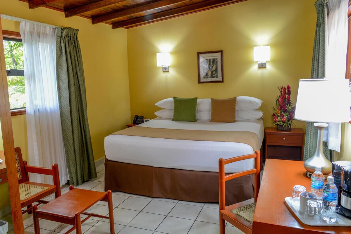 Una habitación de hotel con una cama, dos lámparas de noche, una mesita de noche, una silla, una mesa con un tray de botellas de agua y una ventana con cortinas.