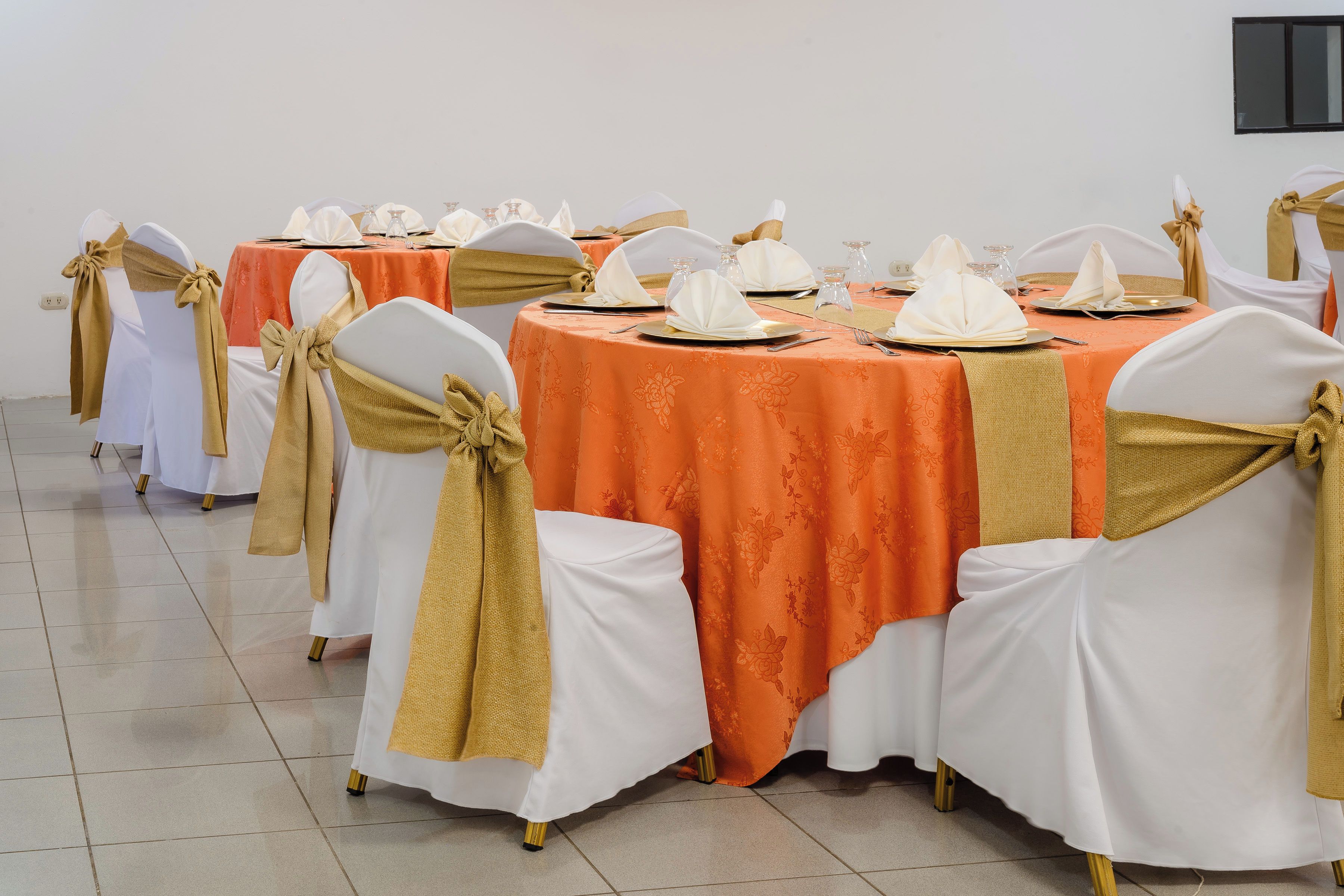 A banquet hall with round tables, white chairs with gold sashes, and orange tablecloths.