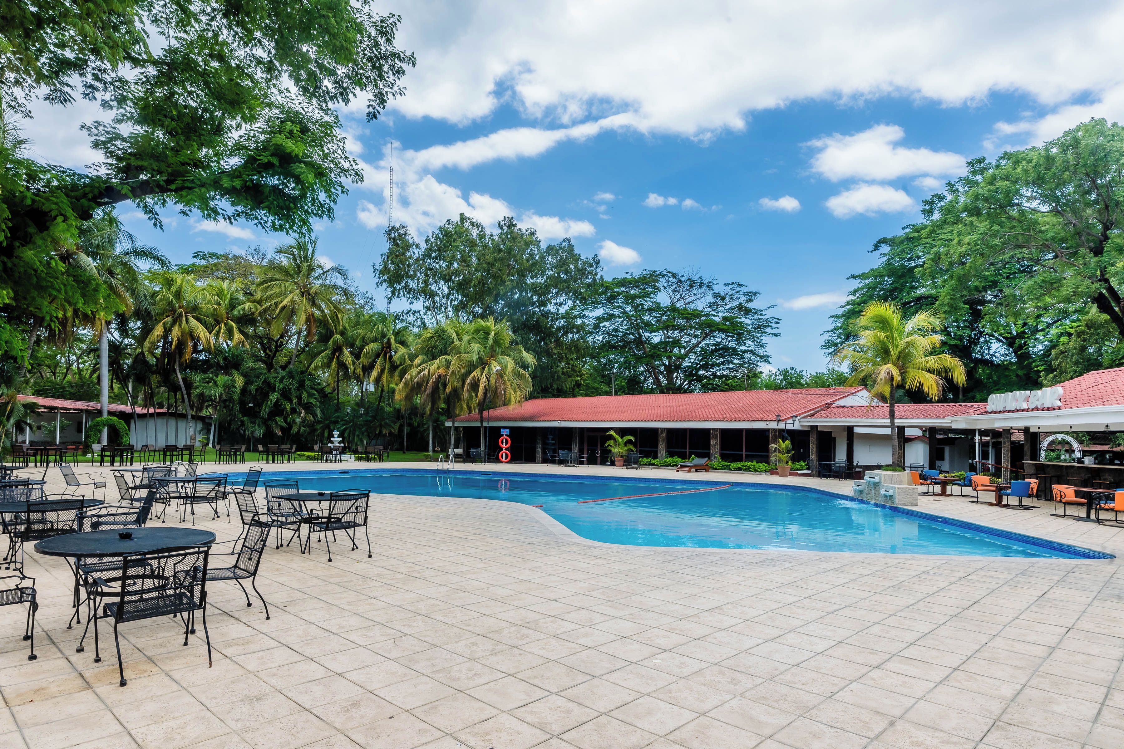 A swimming pool with lounge chairs and tables, surrounded by palm trees and a building with a red roof.