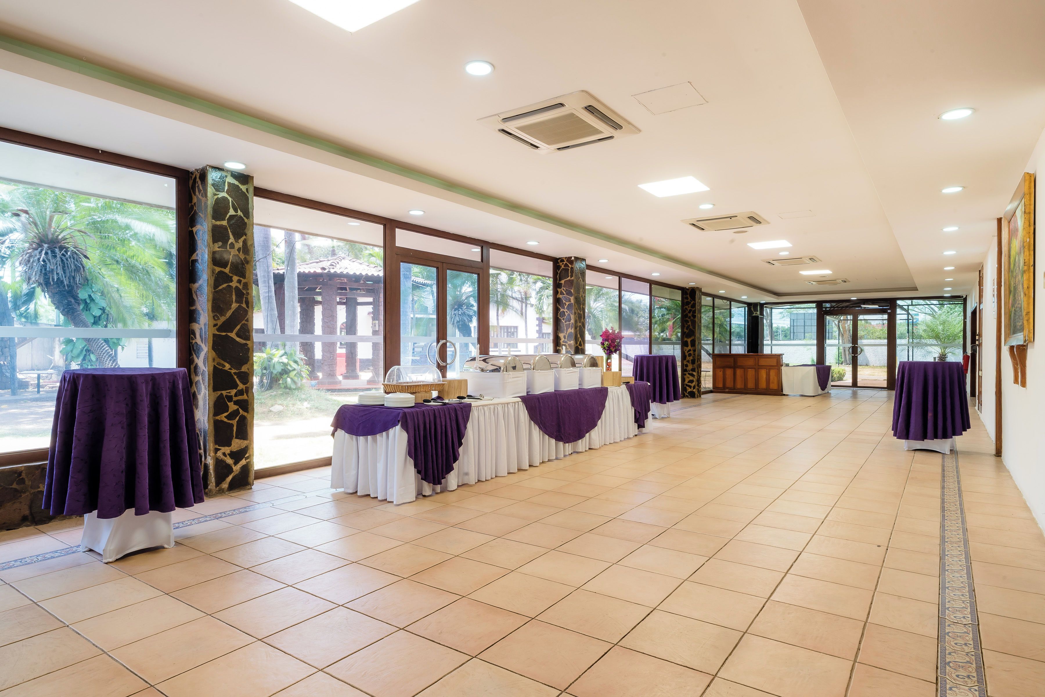 A spacious indoor area with a long table set up for dining, large windows, and a ceiling with recessed lighting.