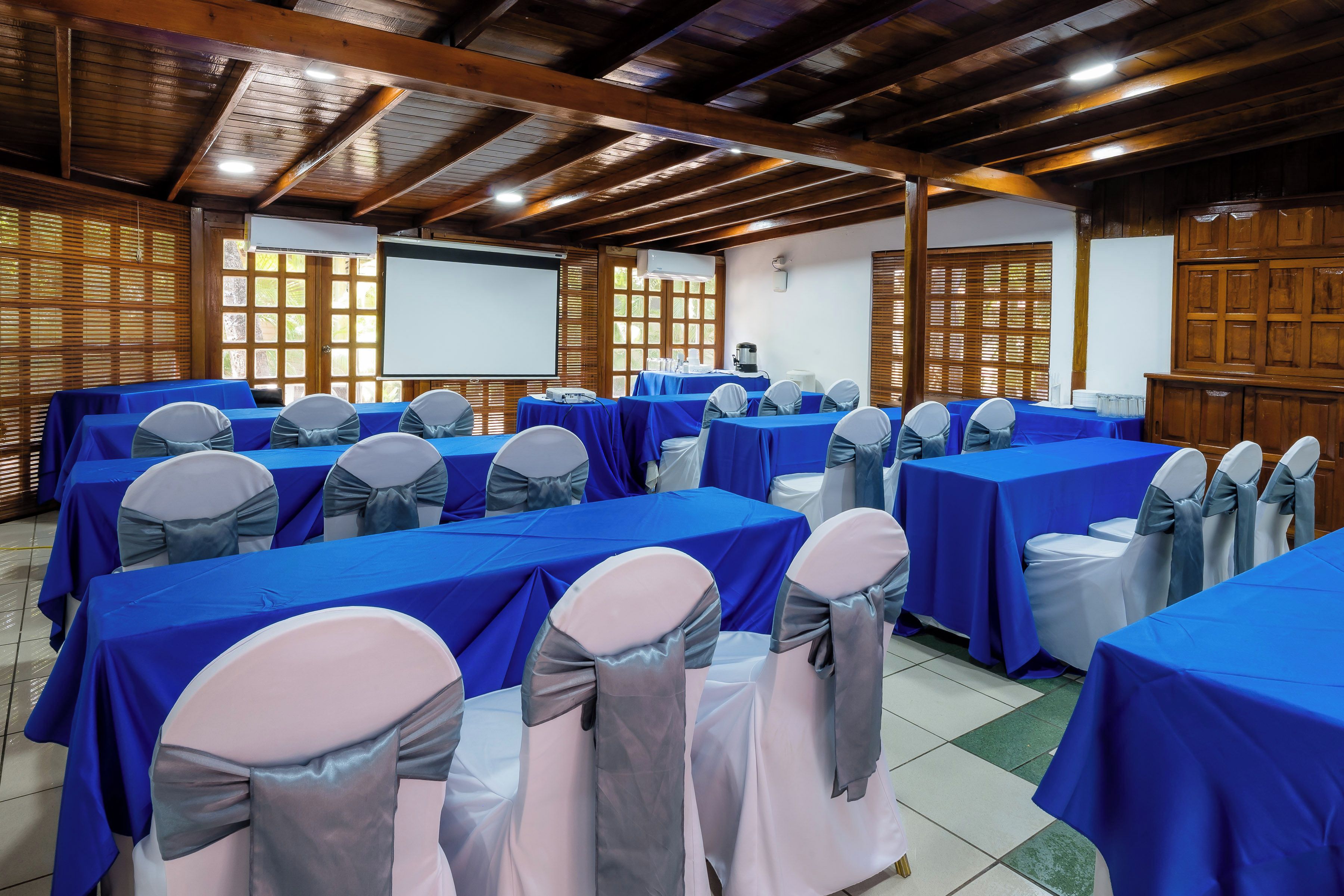 A conference room with multiple tables covered in blue tablecloths, chairs with grey sashes, wooden paneling on the walls, and a projector screen.