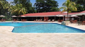 Una piscina con sillas de descanso y sombrillas, rodeada de palmeras y un edificio de techo rojo.