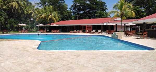 A swimming pool with lounge chairs and umbrellas, surrounded by palm trees and a building with a red roof. - Best Western Las Mercedes Airport (Managua)