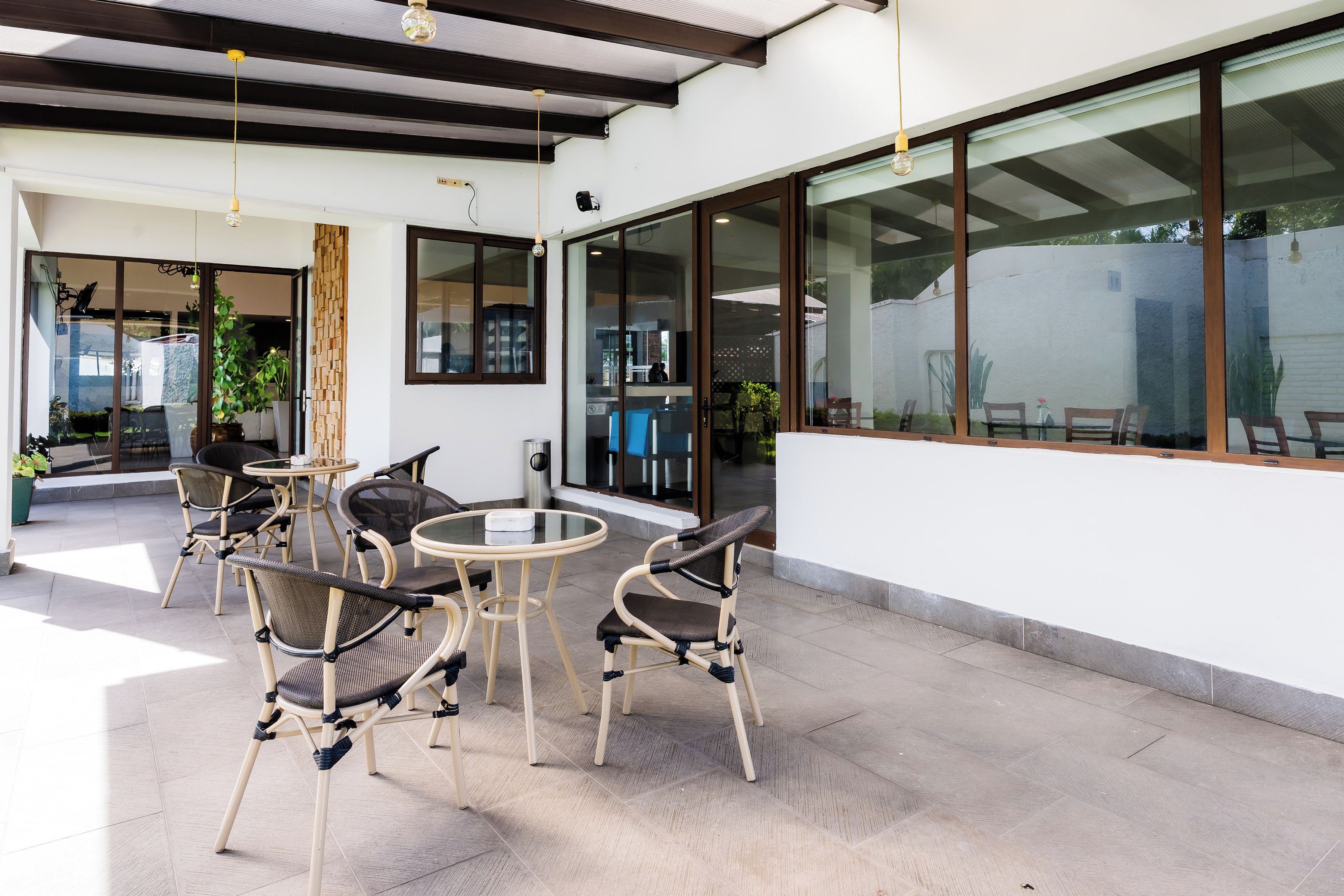 An outdoor seating area with wicker chairs and tables, a glass wall, and a ceiling with wooden beams.