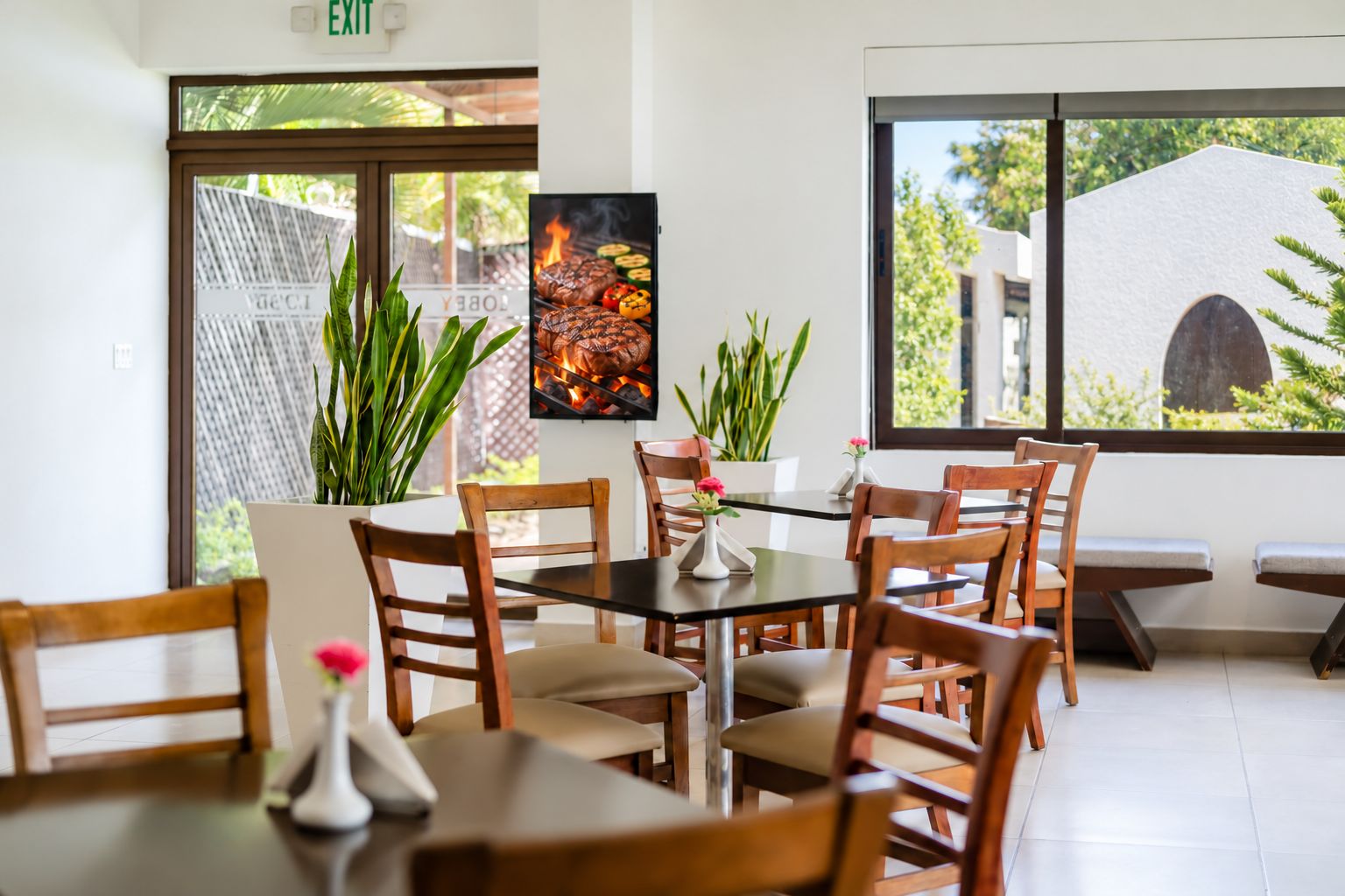 L’intérieur d’un restaurant, avec un espace repas, des chaises en bois, une table agrémentée d’un vase et une grande fenêtre donnant sur un paysage verdoyant.