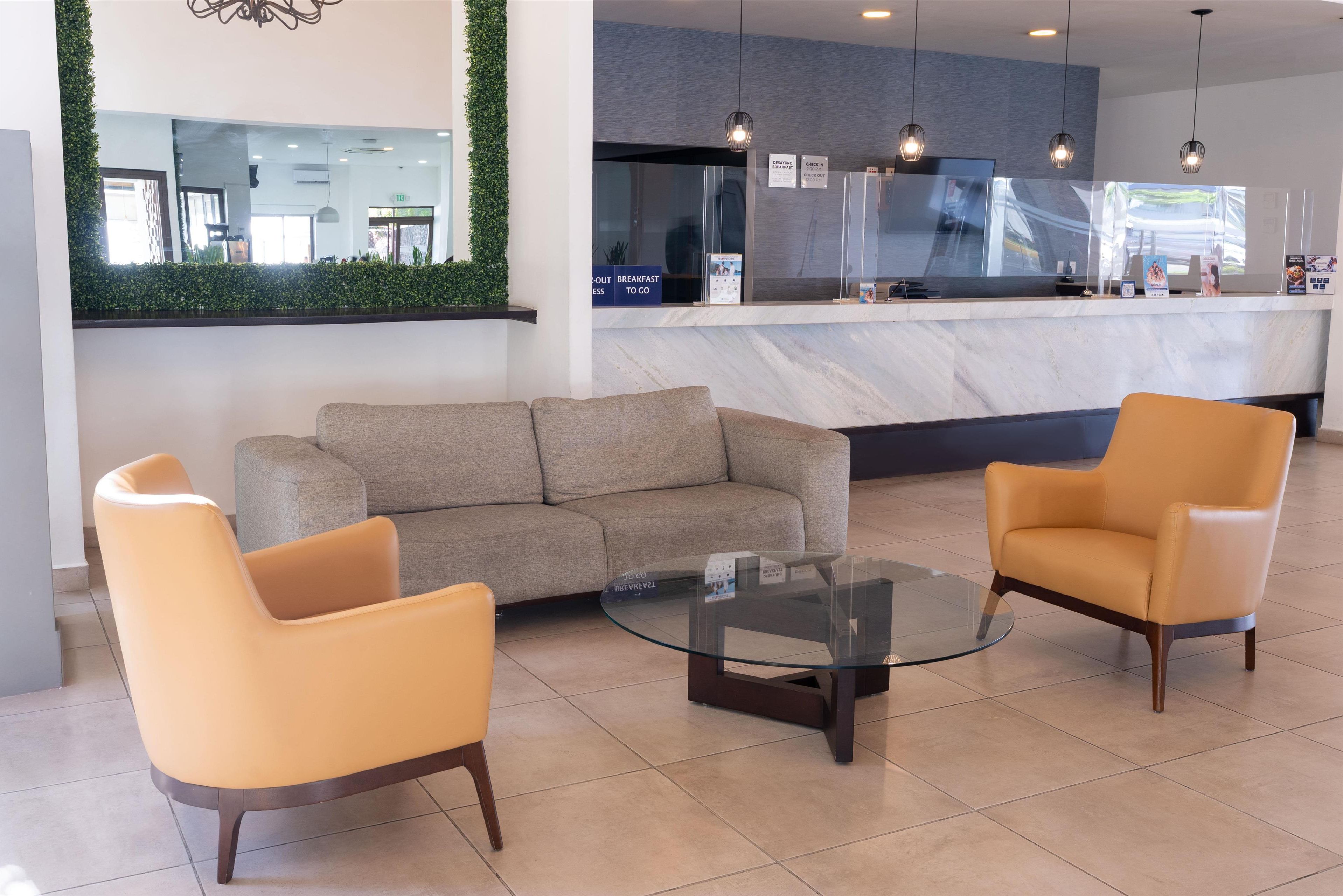 A modern lobby with a sofa, two armchairs, a glass coffee table, and a reception desk with a mirror and greenery.