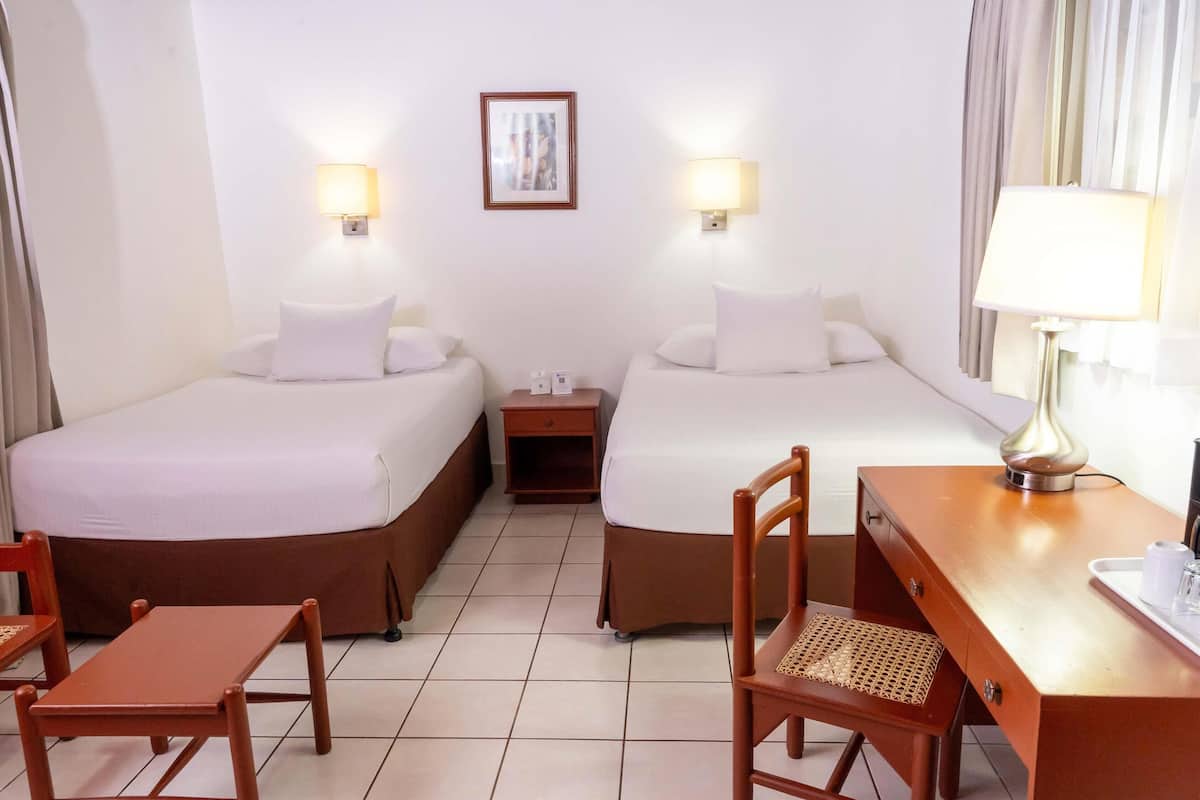 Habitación de hotel con dos camas, un escritorio, una silla, una mesita de noche y una lámpara.