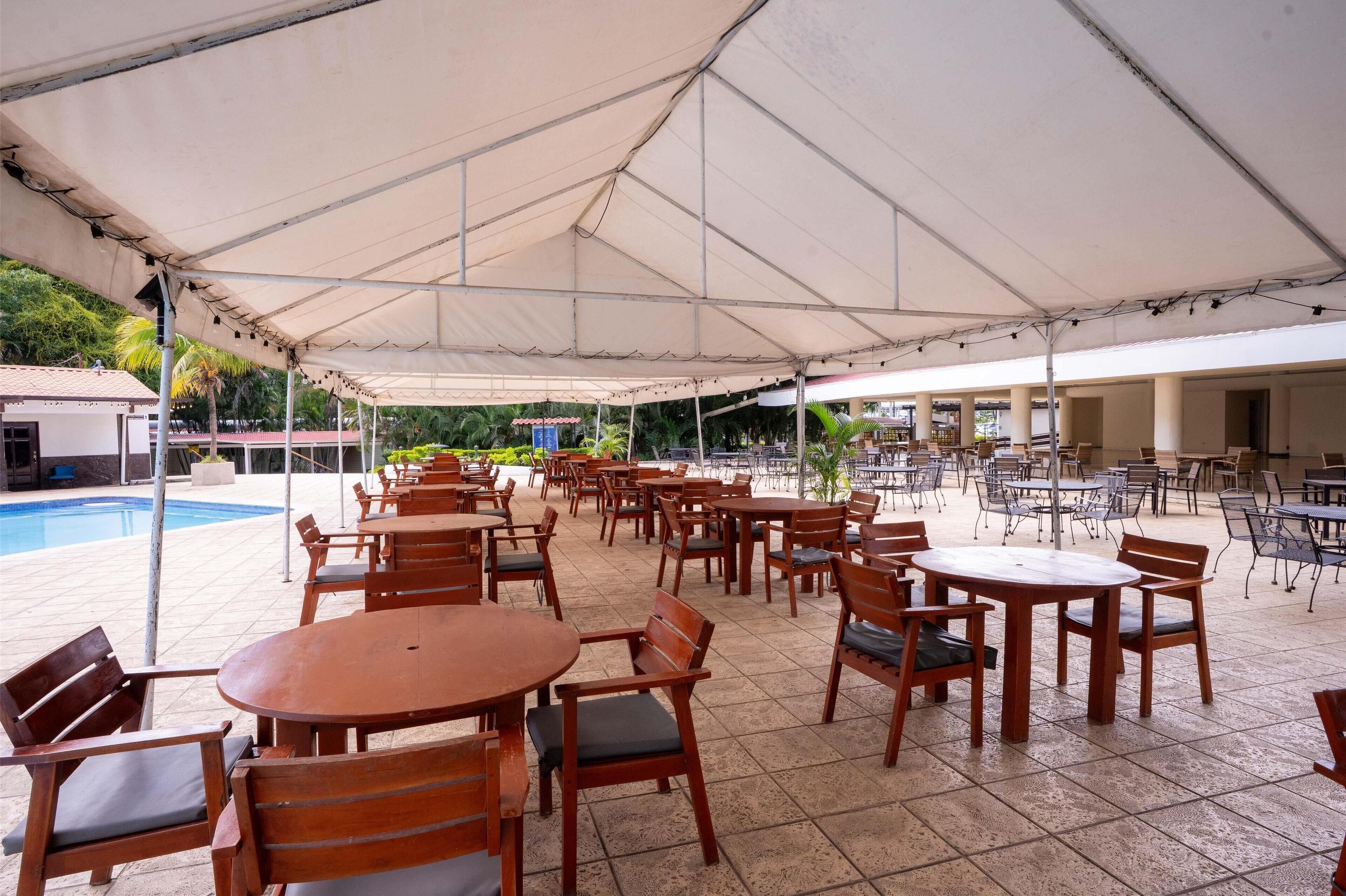 Un área de patio al aire libre con varias mesas y sillas de madera, una piscina al fondo y una sección cubierta con sombrillas.