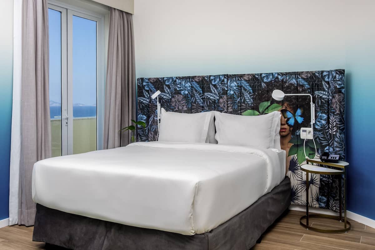 A bedroom with a bed, a bedside table, a lamp, and a view of the sea through a window.