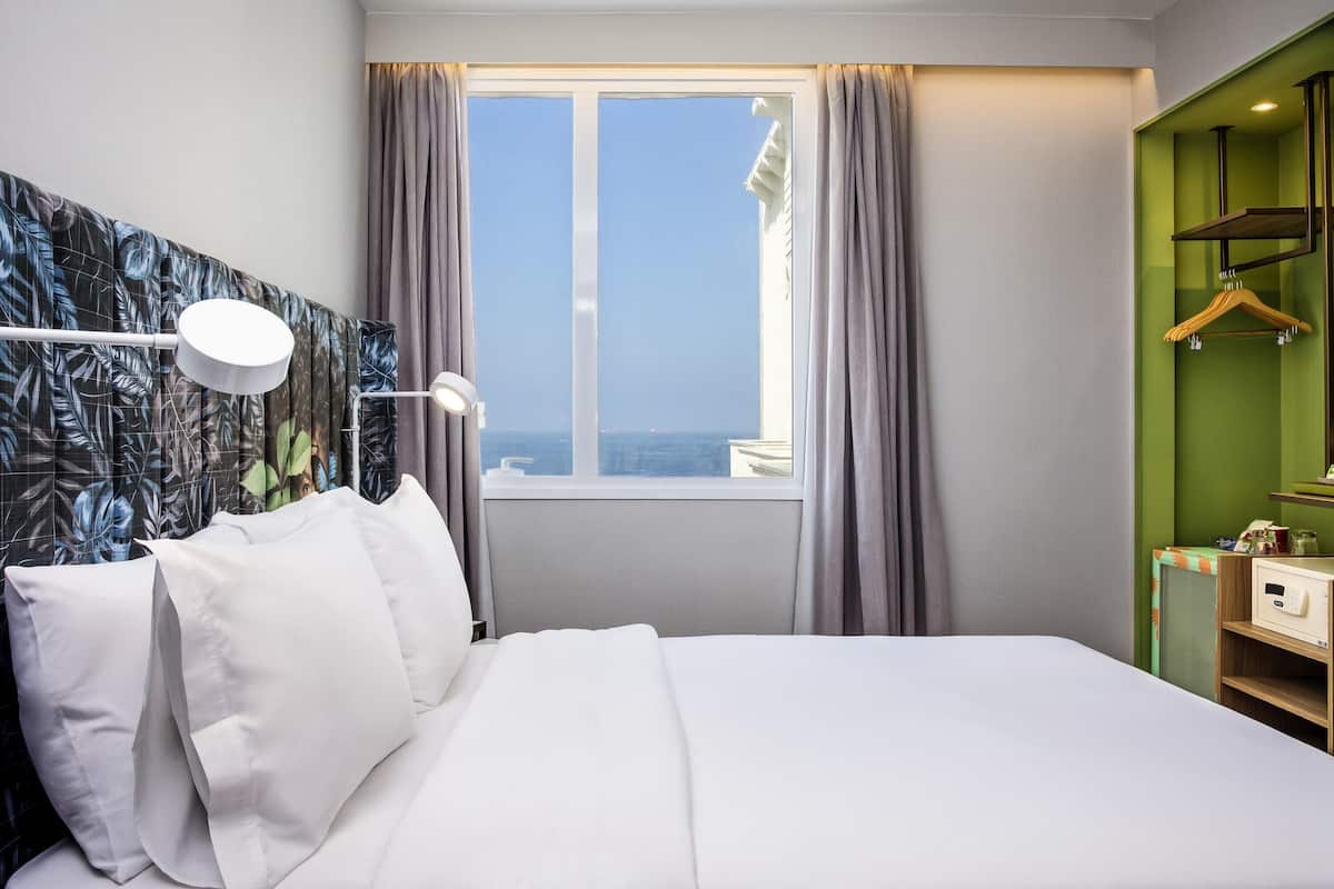A hotel room with a bed, a window with curtains, a bedside lamp, and a green wall with a mirror and shelves.