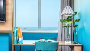 A room with a desk, a chair, a window with a view of the sea, a lamp, and books. - Mercure Rio Boutique Hotel Copacabana (Rio de Janeiro)