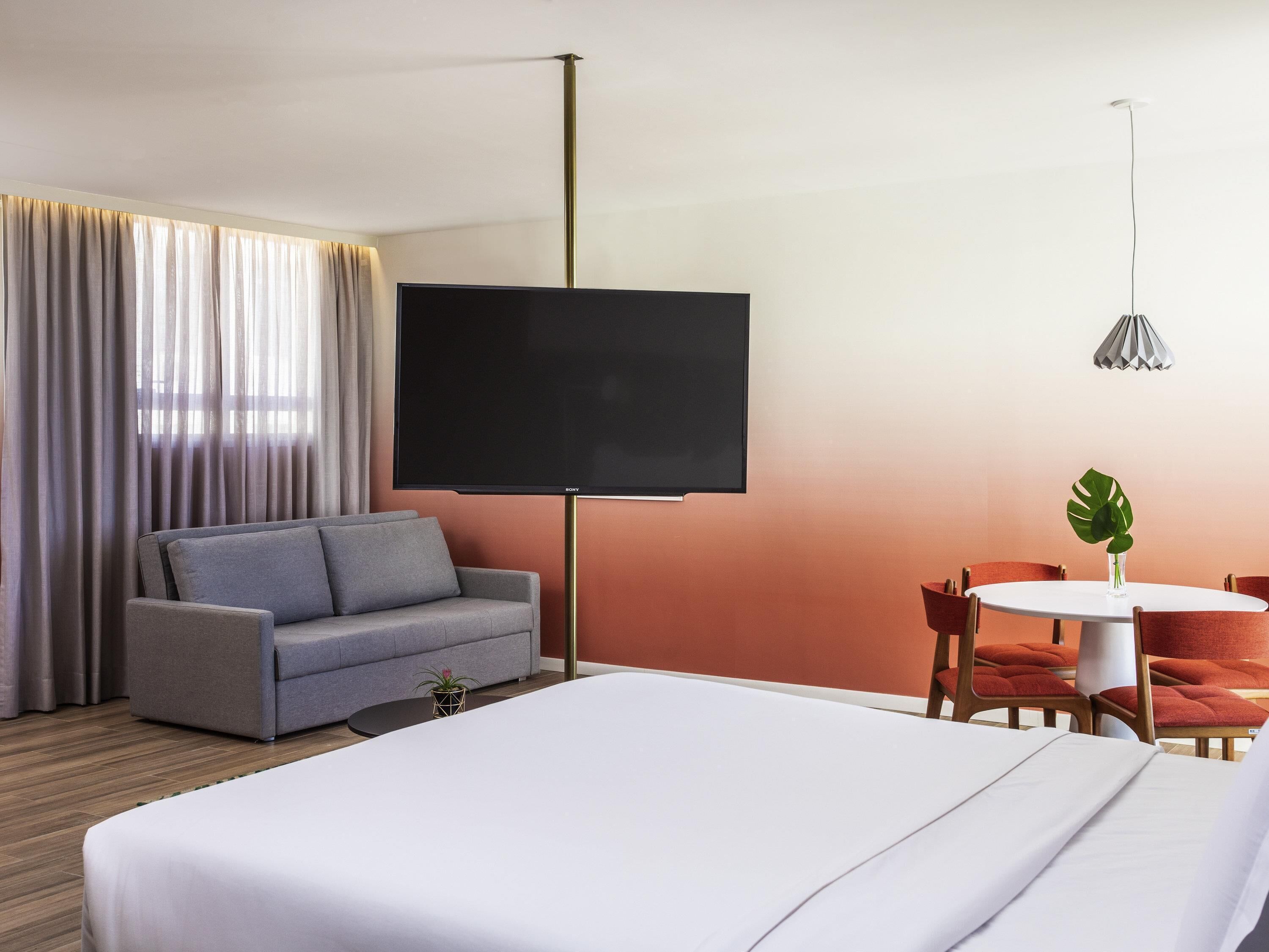 A hotel room with a bed, a sofa, a TV, a dining table with chairs, and a potted plant.