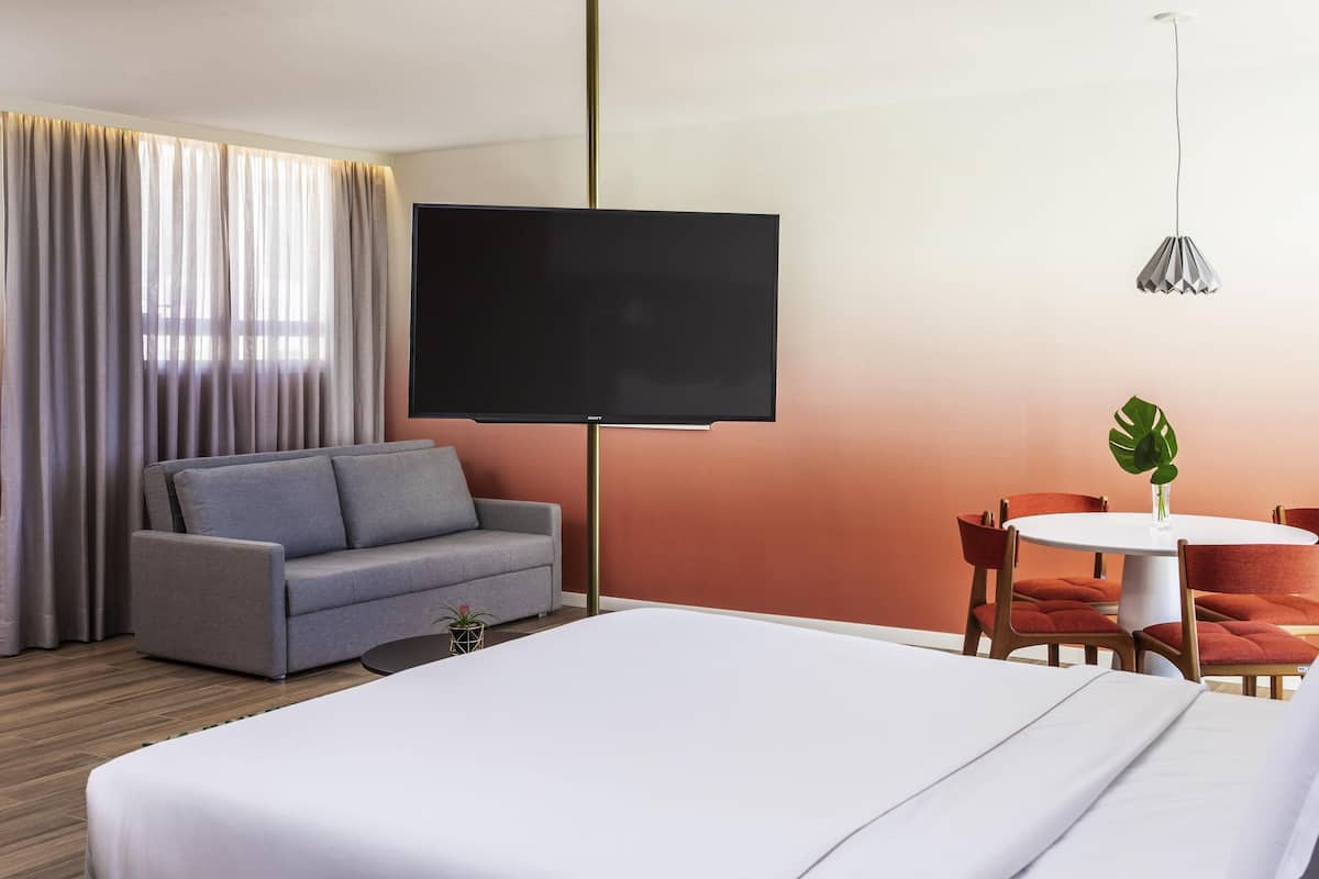 A hotel room with a bed, a sofa, a TV, a dining table with chairs, and a potted plant.