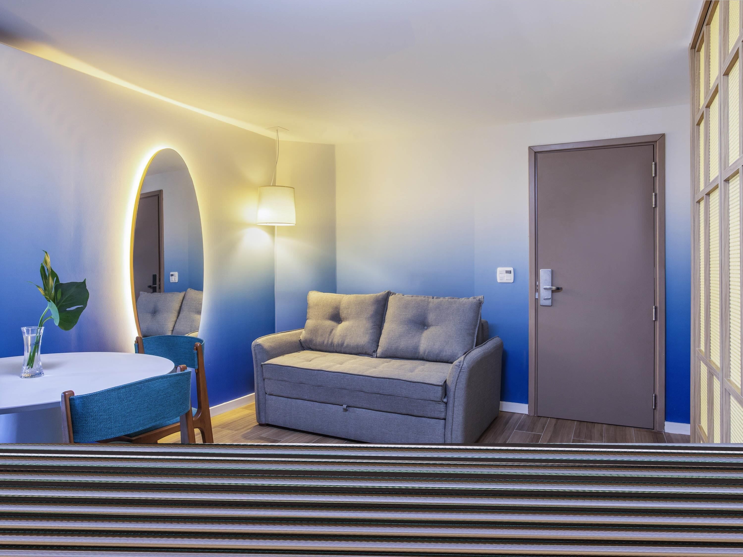 Een kamer met een bank, een ronde tafel, een lamp, een spiegel en een deur.